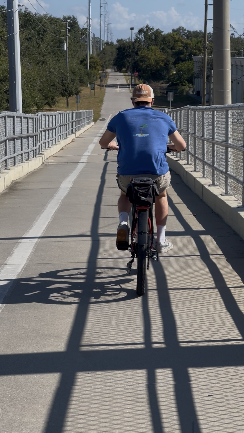 A person riding a bike on a dedicated bike lane with metal railings on both sides, casting shadows on the pavement, under a partly cloudy sky.