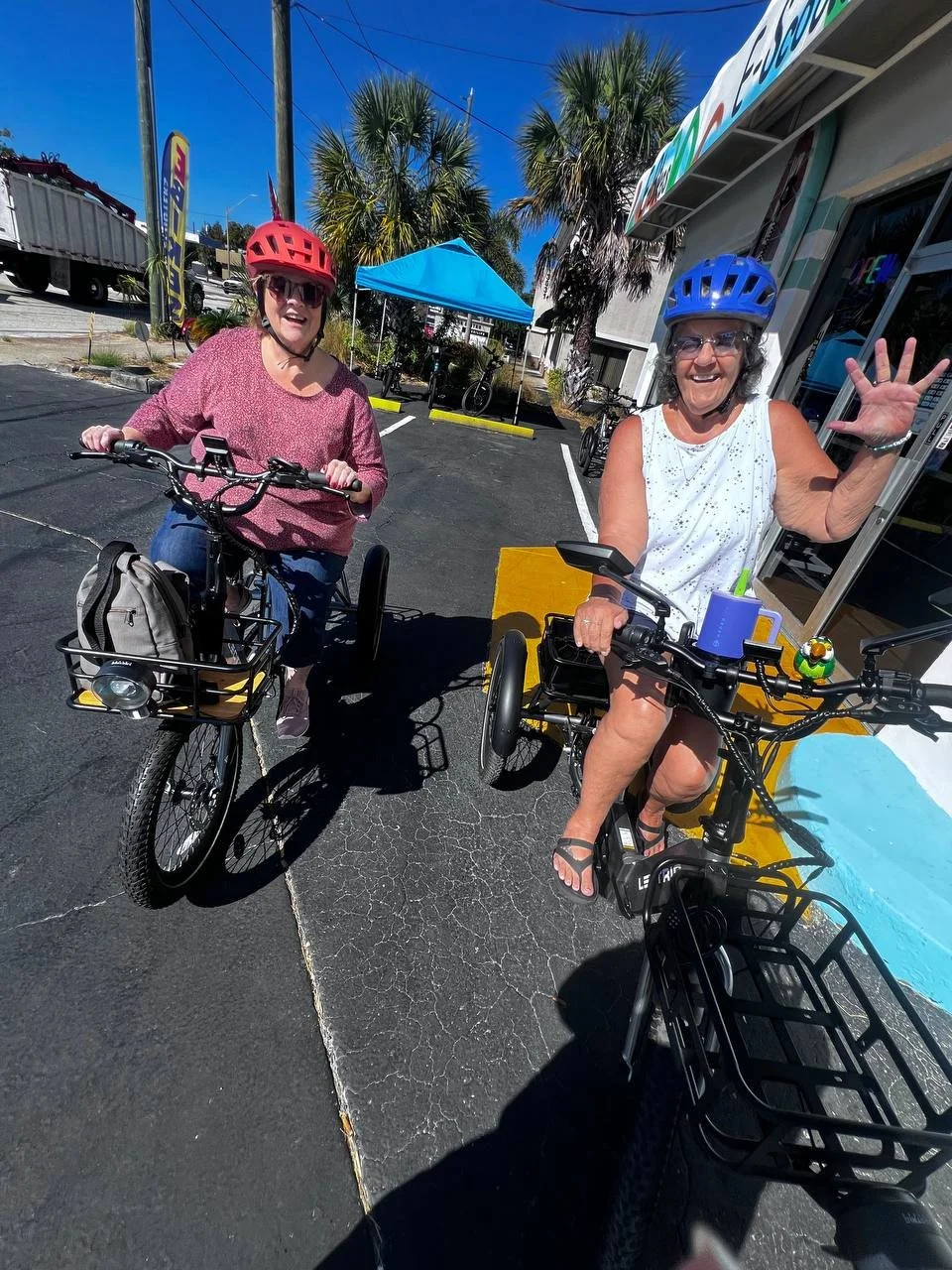 Two women smiling and waving while riding bikes with attached baskets in a parking lot under sunny weather, wearing helmets, with palm trees and a blue canopy in the background.