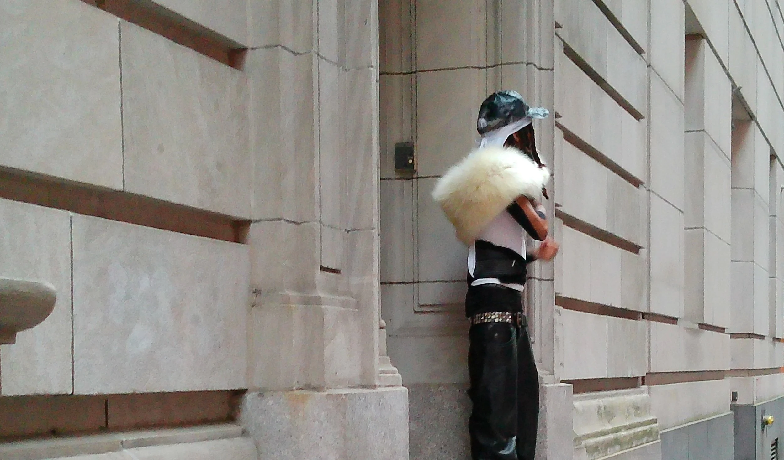 A A man with dreads wearing a camouflage cap, white durag, a vintage yellow fur scarf, and black leather pants stands against a beige stone wall with square windows.