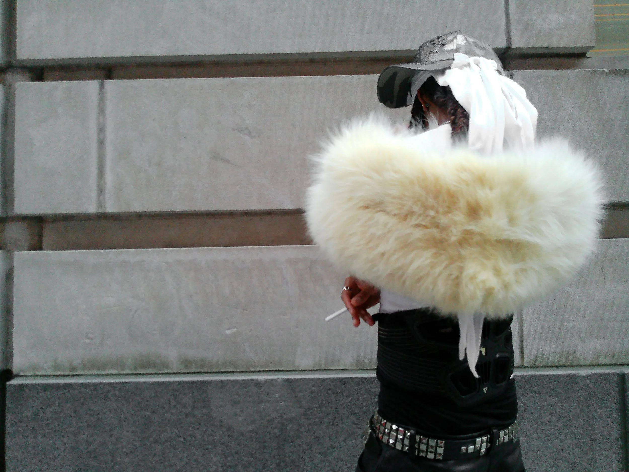 A man with dreads wearing a camouflage cap, white durag, a vintage yellow fur scarf, and spike-studded belt in front of a New York building on the street. Industrial attitude, chic outfit.