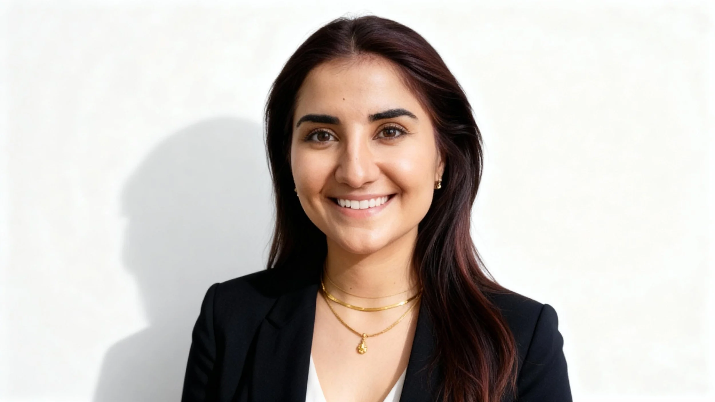 A smiling young woman with long dark hair, wearing a black blazer, white top, and gold jewelry, standing against a white background.
