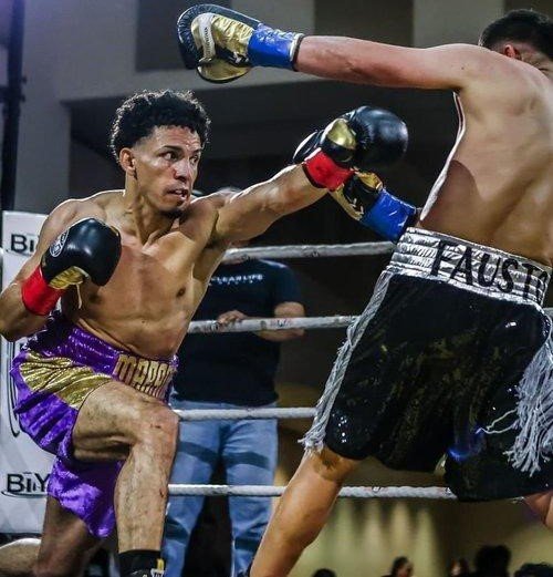 Two boxers in action in a ring, one throwing a punch towards the other. The boxer on the left is wearing purple and gold shorts, and the boxer on the right has "FAUST" on their shorts. The scene captures the intensity and motion of a boxing match.