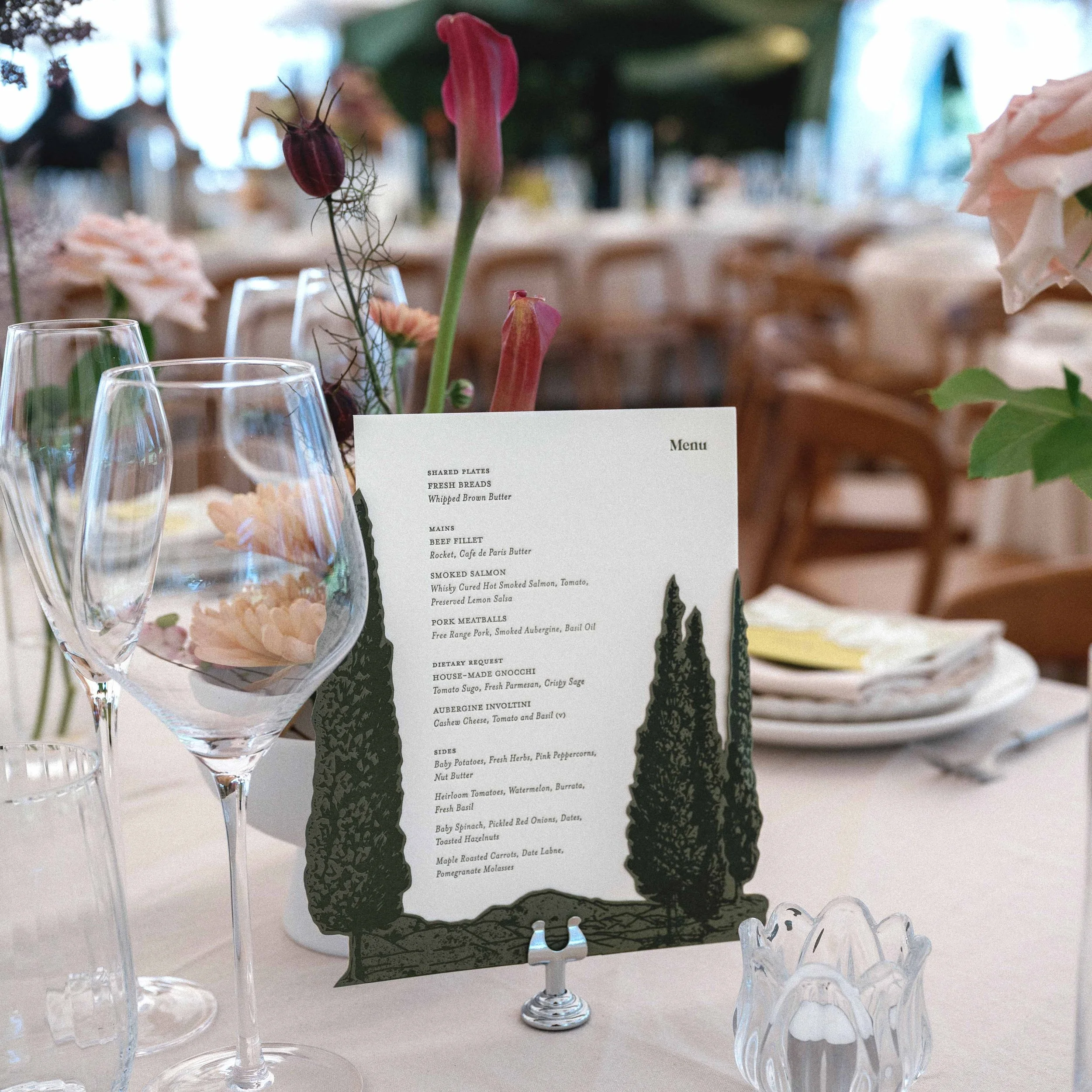 Greer and Cohen’s die-cut letterpress dinner menu with layered conifers, displayed among flowers and glassware on their wedding table.