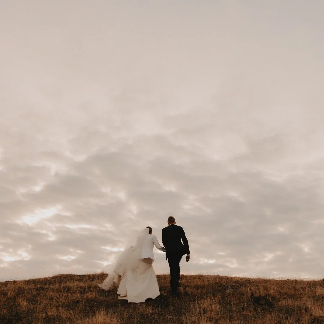 Brontë and Simon walking through the tussock-covered clifftops at Boomrock on their wedding day.
