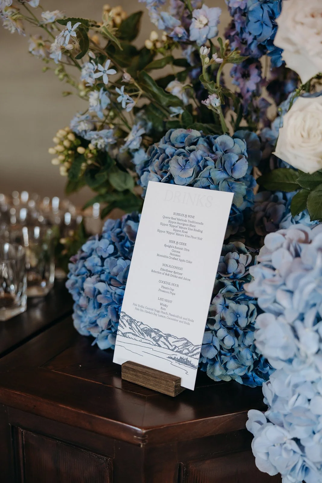Letterpress drinks menu featuring a hand-drawn landscape illustration displayed beside blue floral arrangements at Rippon, Wānaka.