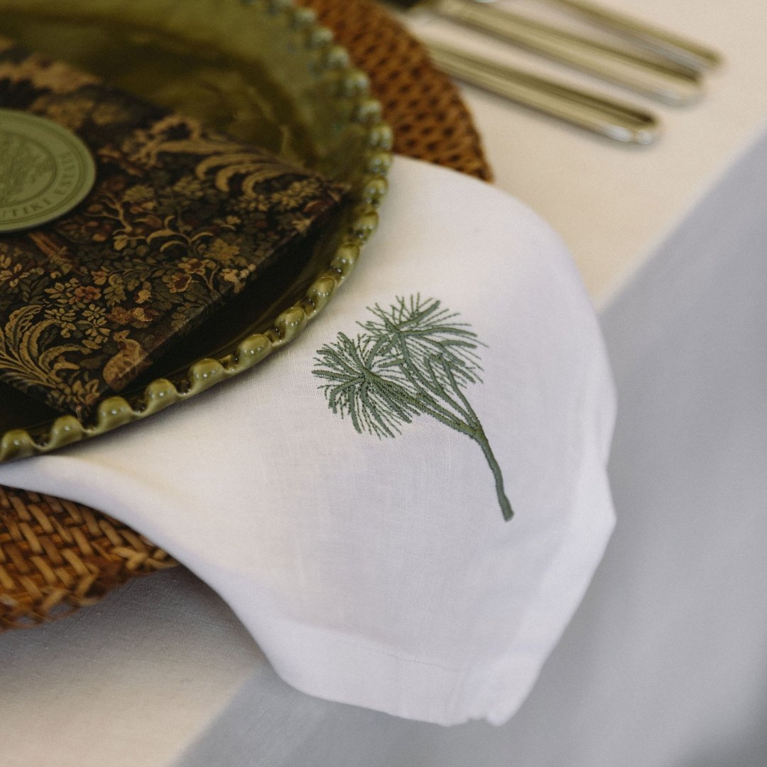 Embroidered tī kōuka illustration on a white wedding napkin under a menu with tapestry artwork at Putiki Estate.