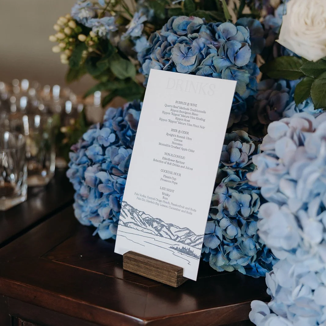 Izzy and Hayden’s letterpress drinks menu with Rippon landscape illustration, displayed among blue hydrangeas at their Wānaka wedding.