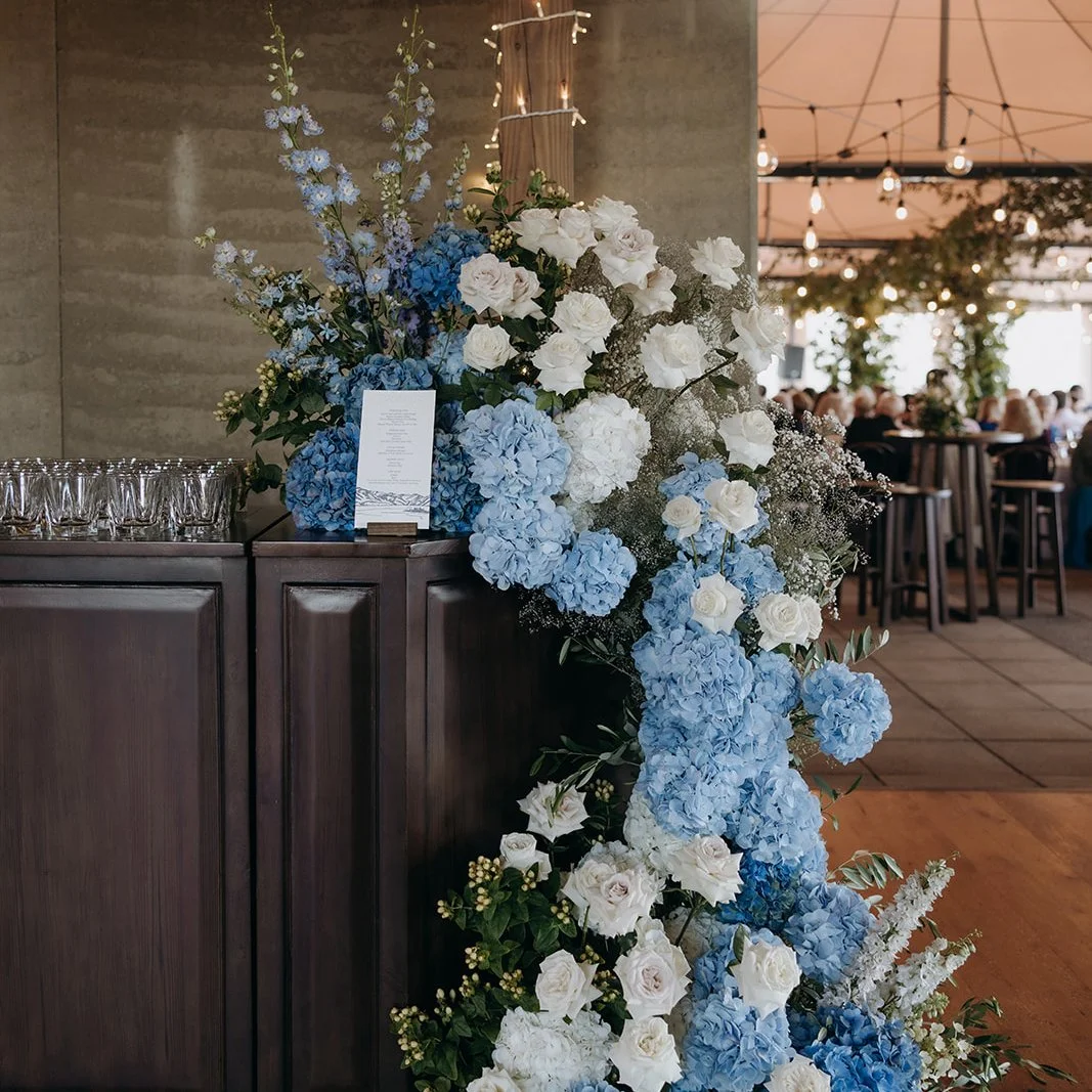 Izzy and Hayden’s letterpress wedding drinks menu displayed amongst blue and white florals at Rippon, Wānaka.