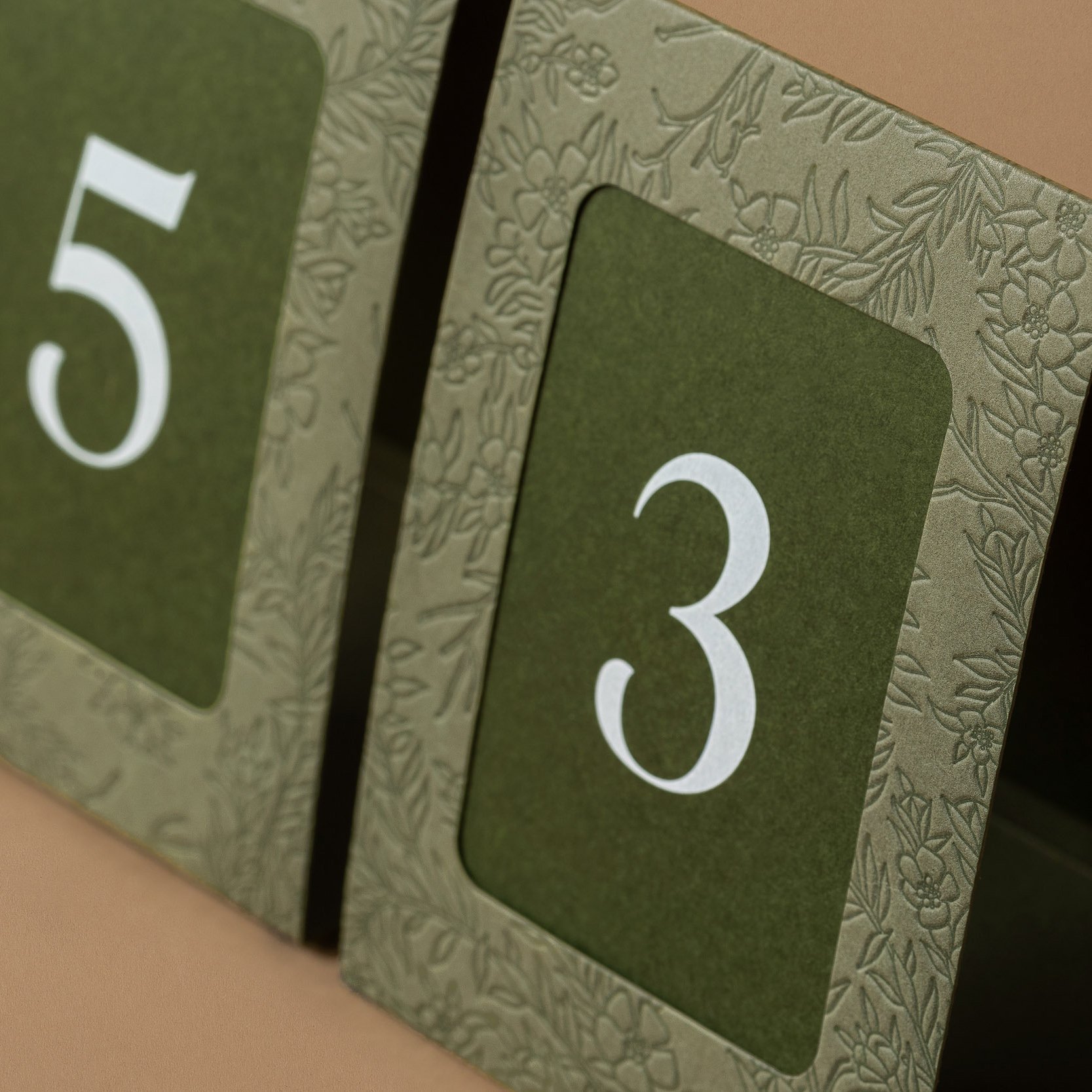 Green wedding table numbers with die-cut frames featuring embossed mānuka artwork and white printed numerals.