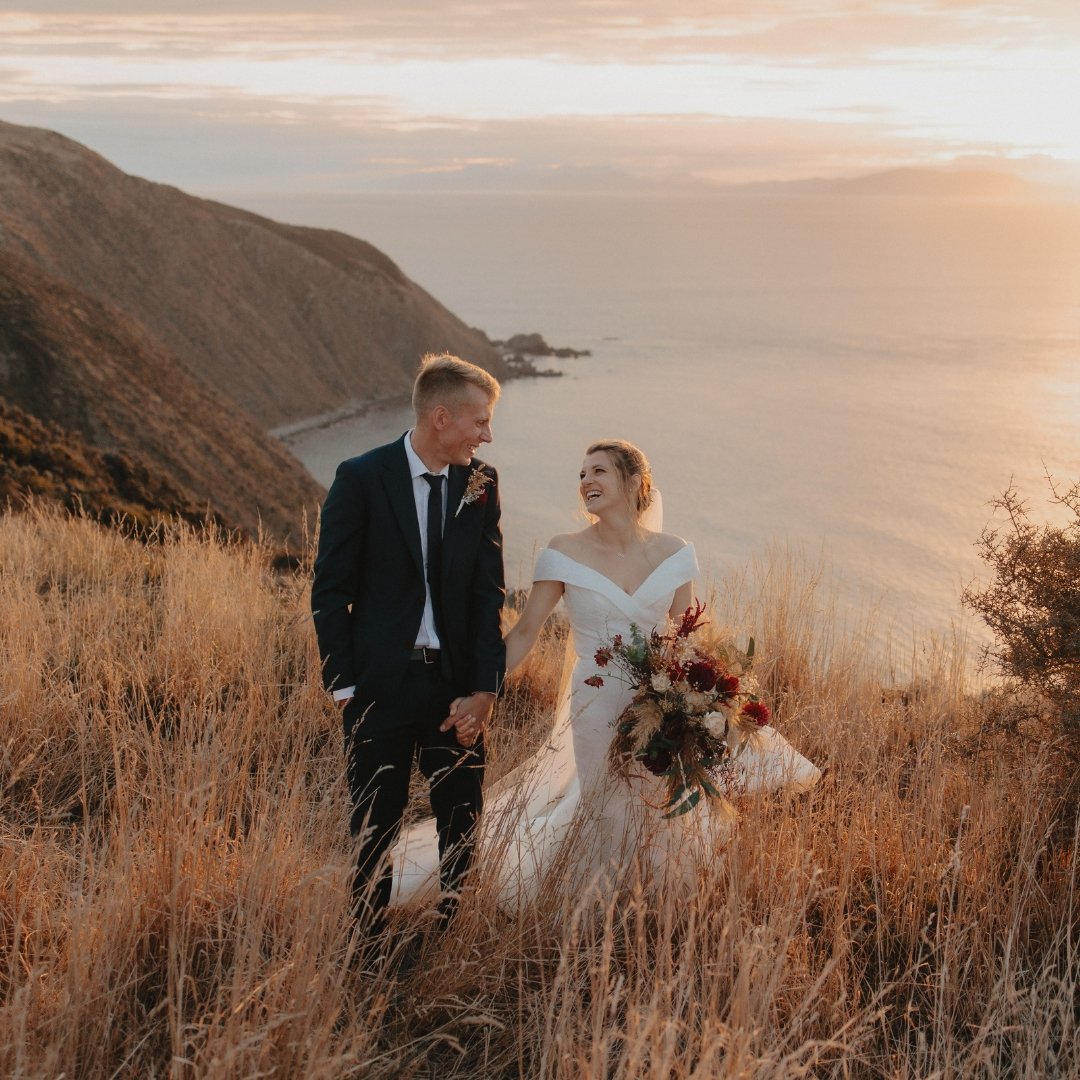 Brontë and Simon walking through tussock grass at Boomrock, with the coastline and ocean behind them at sunset.