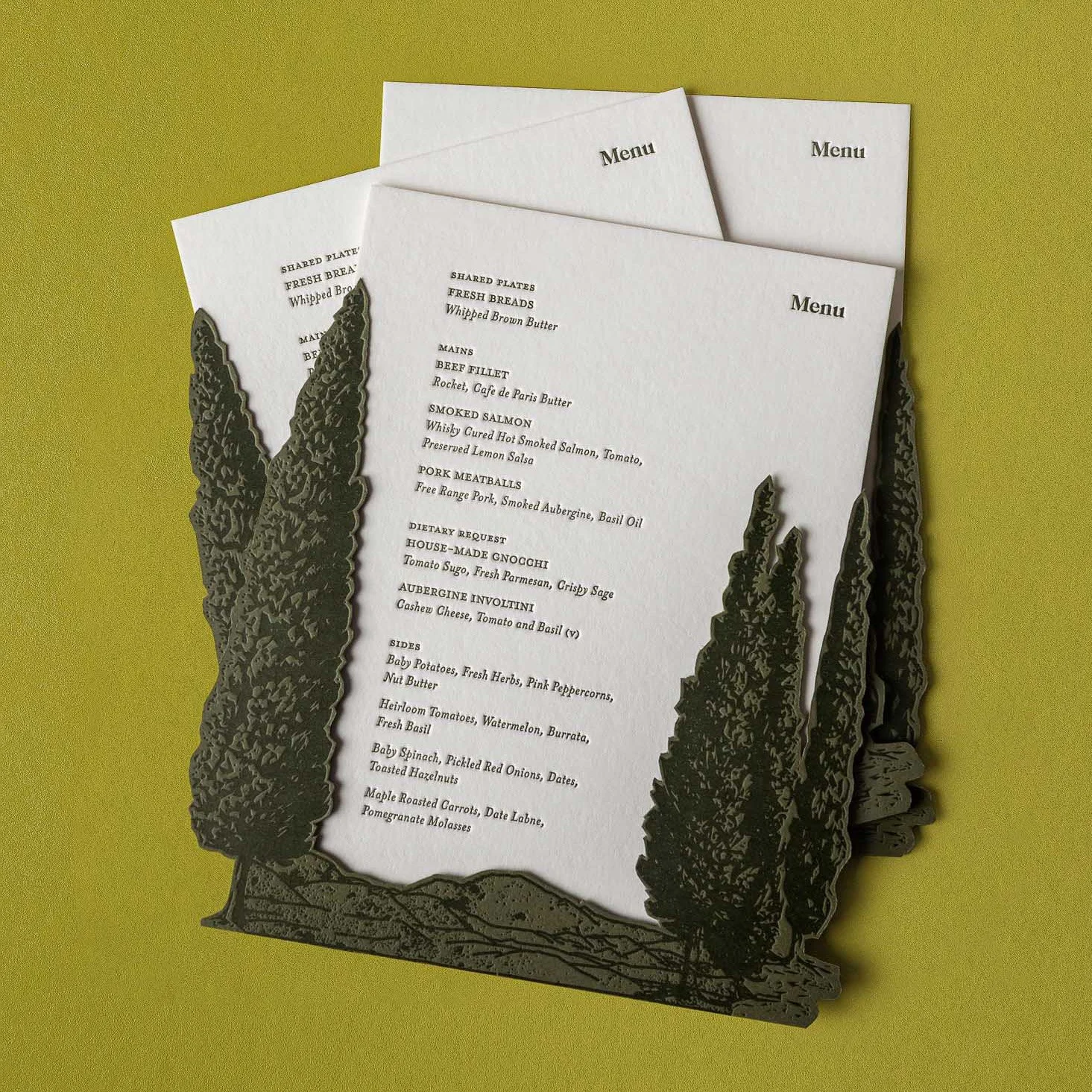 Greer and Cohen’s wedding dinner menus layered with die-cut letterpress conifer artwork and an illustration of Mount Starveall.