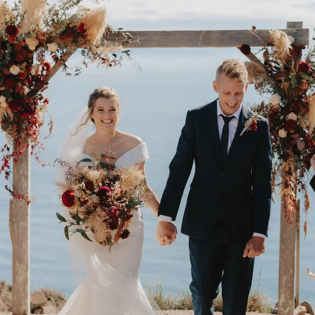 Brontë and Simon beneath the flower-covered ceremony arch at their Boomrock wedding, with the ocean beyond.