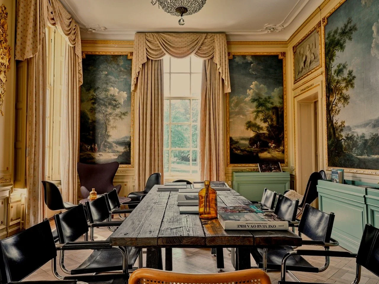 Een vergaderruimte met een grote houten tafel, zwarte stoelen, muurkunst met landschapstafereel, grote ramen met gordijnen, een groene kast, en decoratieve plafonddetails.