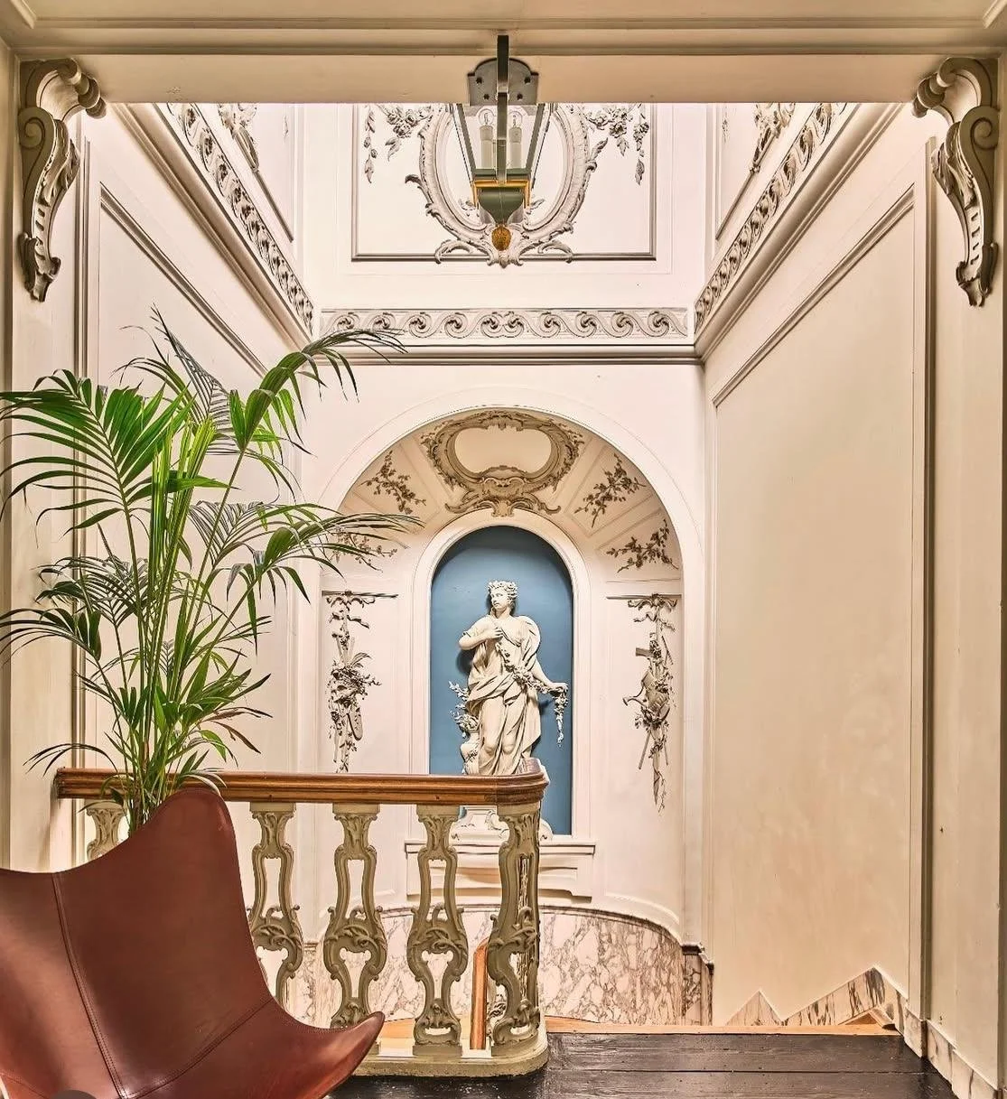 Interieur met een marmeren trap, een houten balustrade, een beeldhouwwerk van een vrouw in een nis, een grote groene plant, en een marmeren vloer.