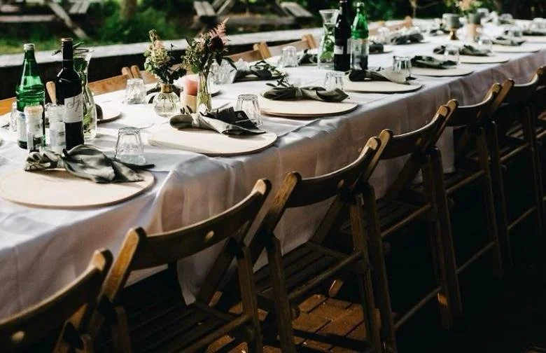 Tafelschikking met witte doek, wijnglazen, wijnflessen, bestek en bloemen op een lange tafel, klaar voor een diner of feest.