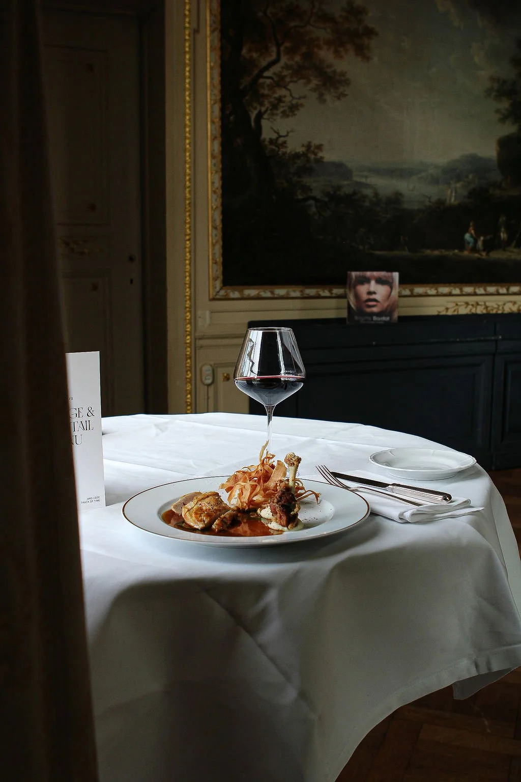 Een gedekte tafel met een bord vol eten, een glas rode wijn en bestek in een elegante kamer met een groot landschapsschilderij op de achtergrond.