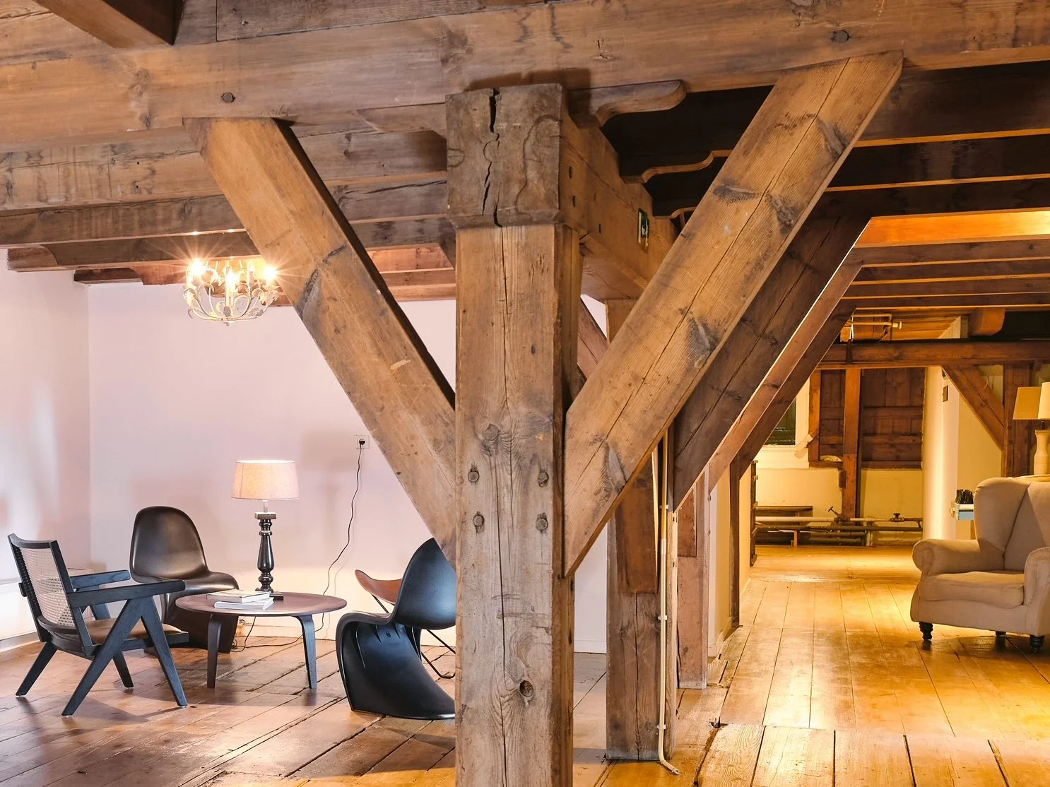 Interieur van een kamer met houten vloer en houten plafond, met meubels zoals een zwarte stoel, een bijzettafel met boeken en een tafellamp, en een beige bank. In het midden een grote houten steunbalk en truss.