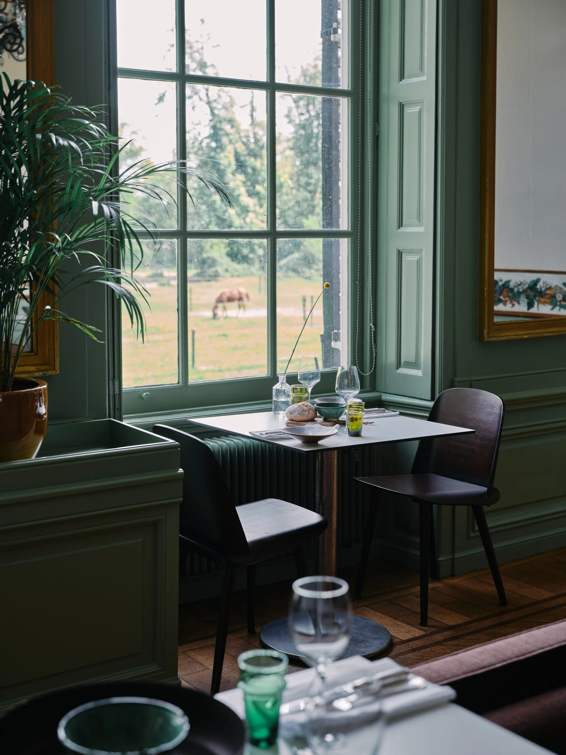 Een binnenkant van een gezellige eetruimte met een grote groene raam met uitzicht op een veld met een paard.