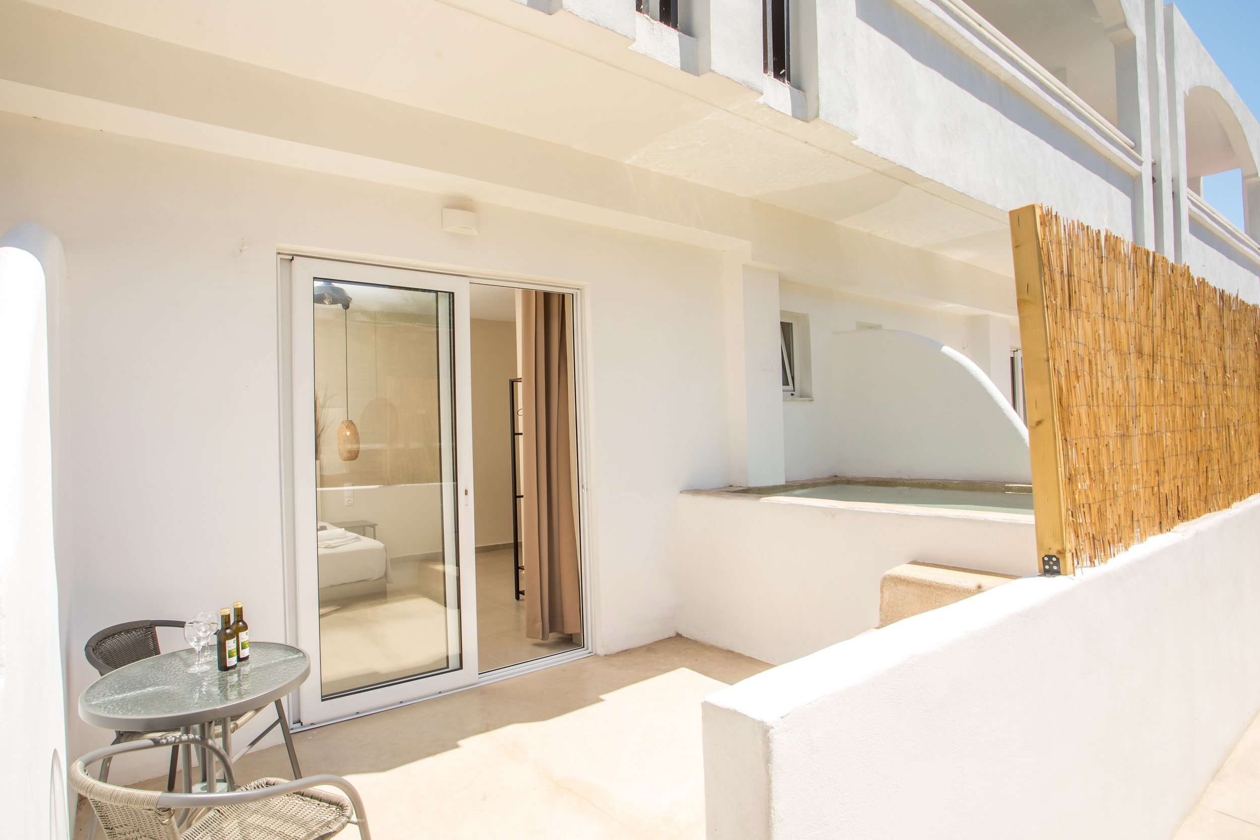 Exterior view of a white Mediterranean-style building with a balcony, sliding glass door leading to a bedroom, a small round table with two wine bottles and glasses, and beige curtains inside the bedroom.