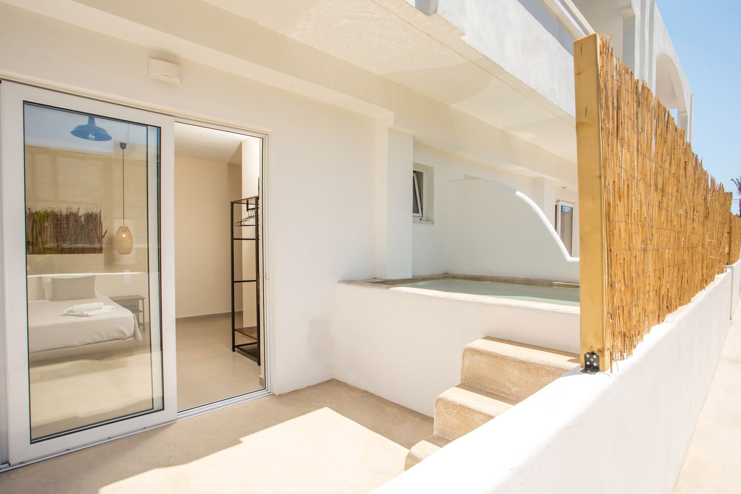 Outdoor patio area with sliding glass door leading to bedroom, small built-in hot tub, and a bamboo privacy screen on a sunny day.