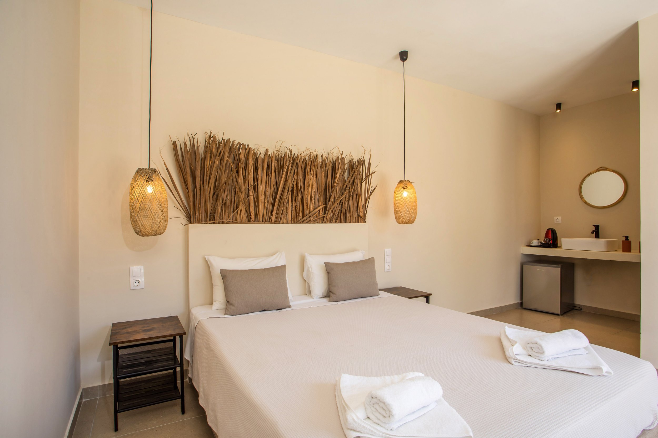 Hotel room with a double bed, beige bedding, two gray pillows, two bedside tables, decorative hanging lights, a wall art made of reeds above the bed, a small sink area with mirror, mini fridge, coffee maker, and towels.
