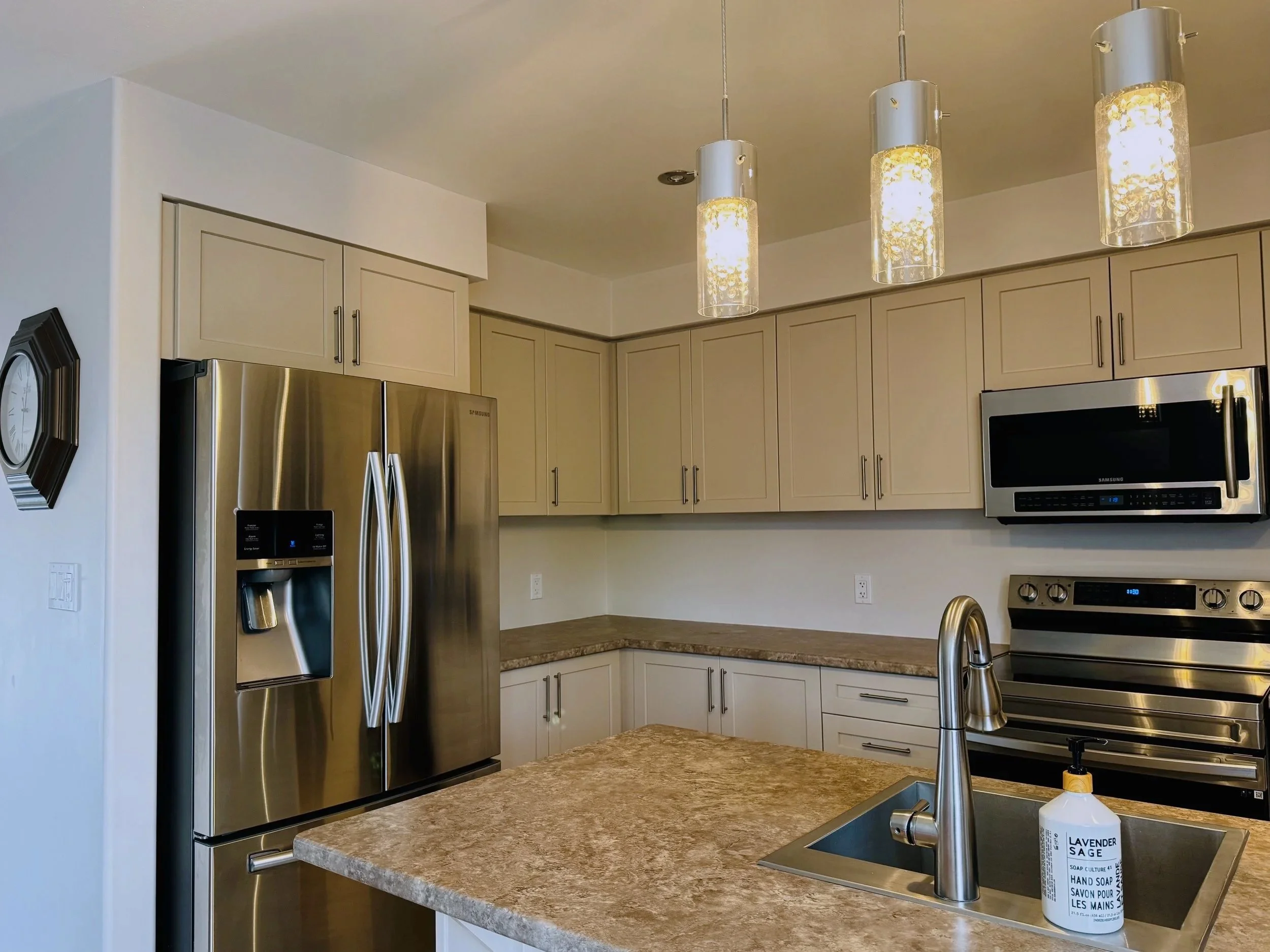 Modern kitchen with stainless steel refrigerator, microwave, oven, beige cabinets, granite countertops, and a small sink with lavender hand soap on the island.