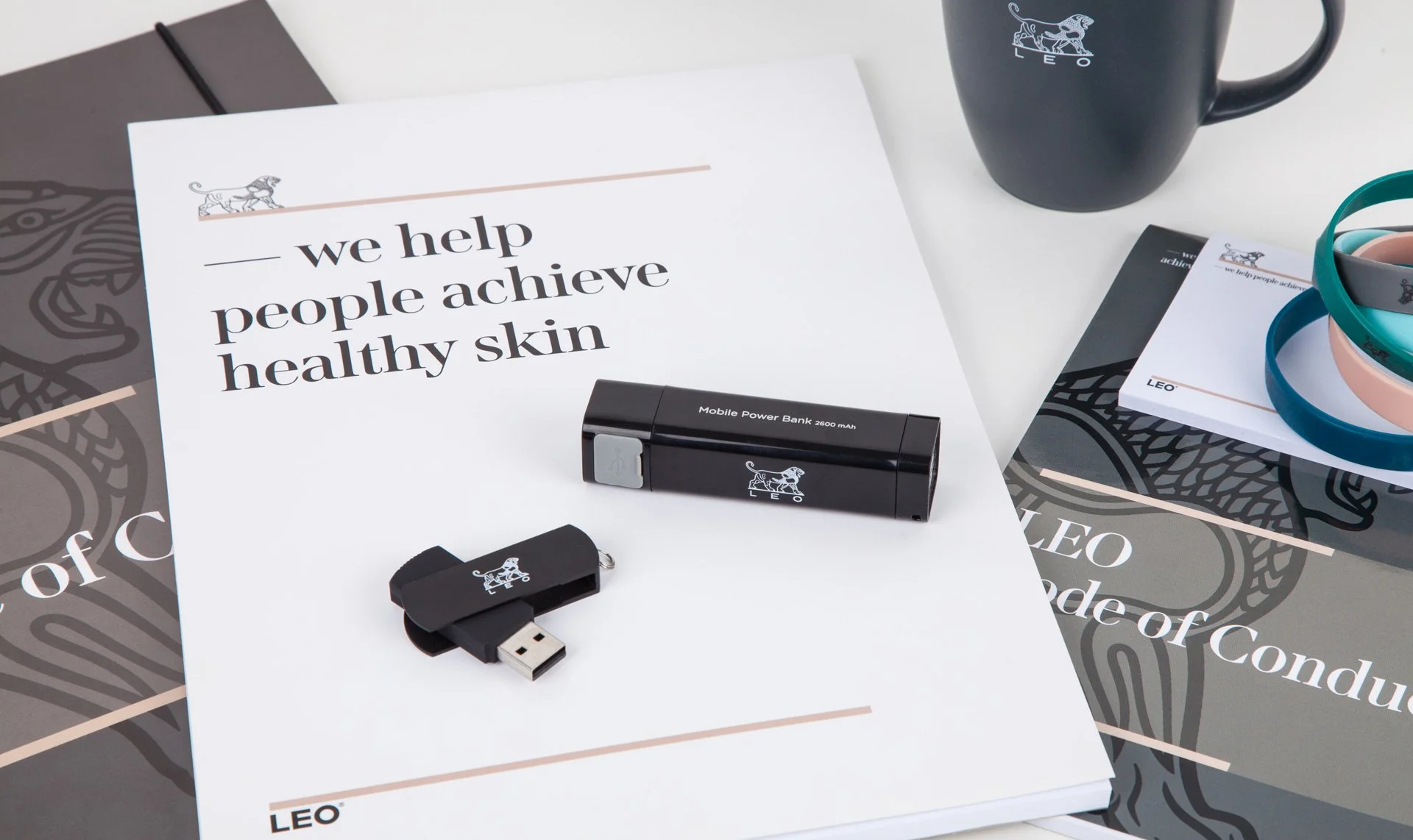Assorted branded items including a black USB drive, a mobile power bank, documents, a mug, and wristbands on a desk.