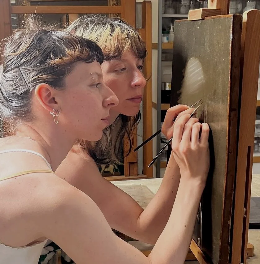 Sisters retouch a small canvas painting using fine brushes, in a conservation studio setting.