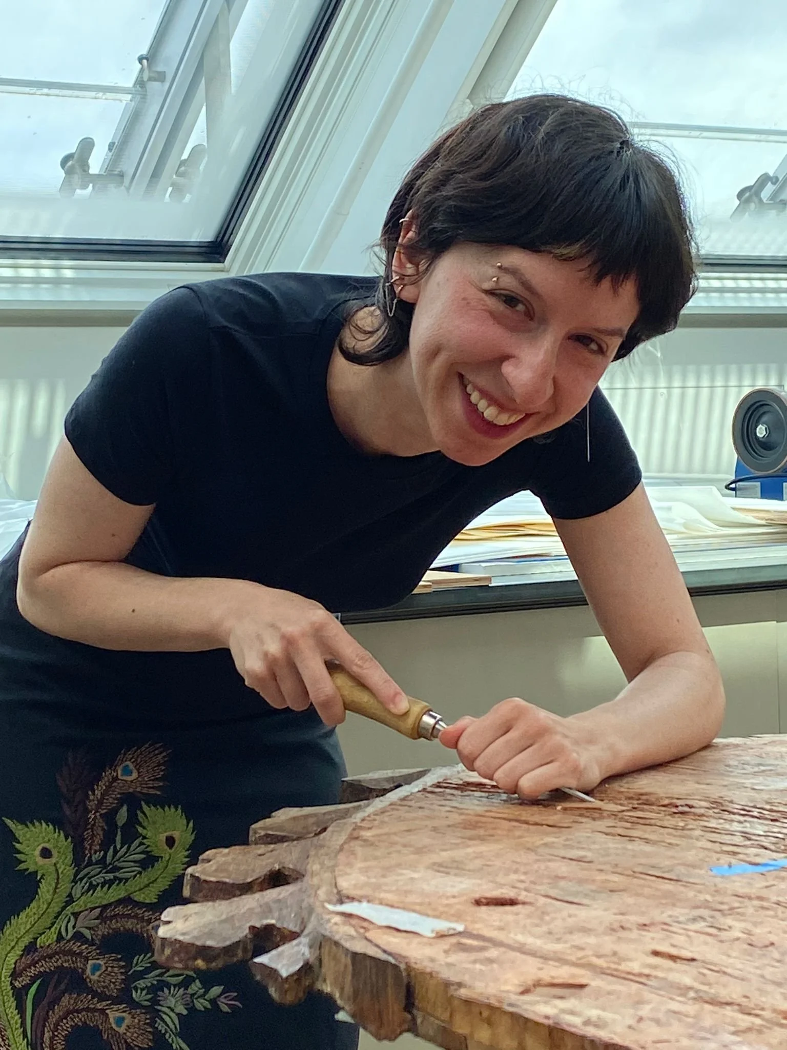 A person with short dark hair, smiling, working on the back of a panel painting with a chisel in a brightly lit studio.