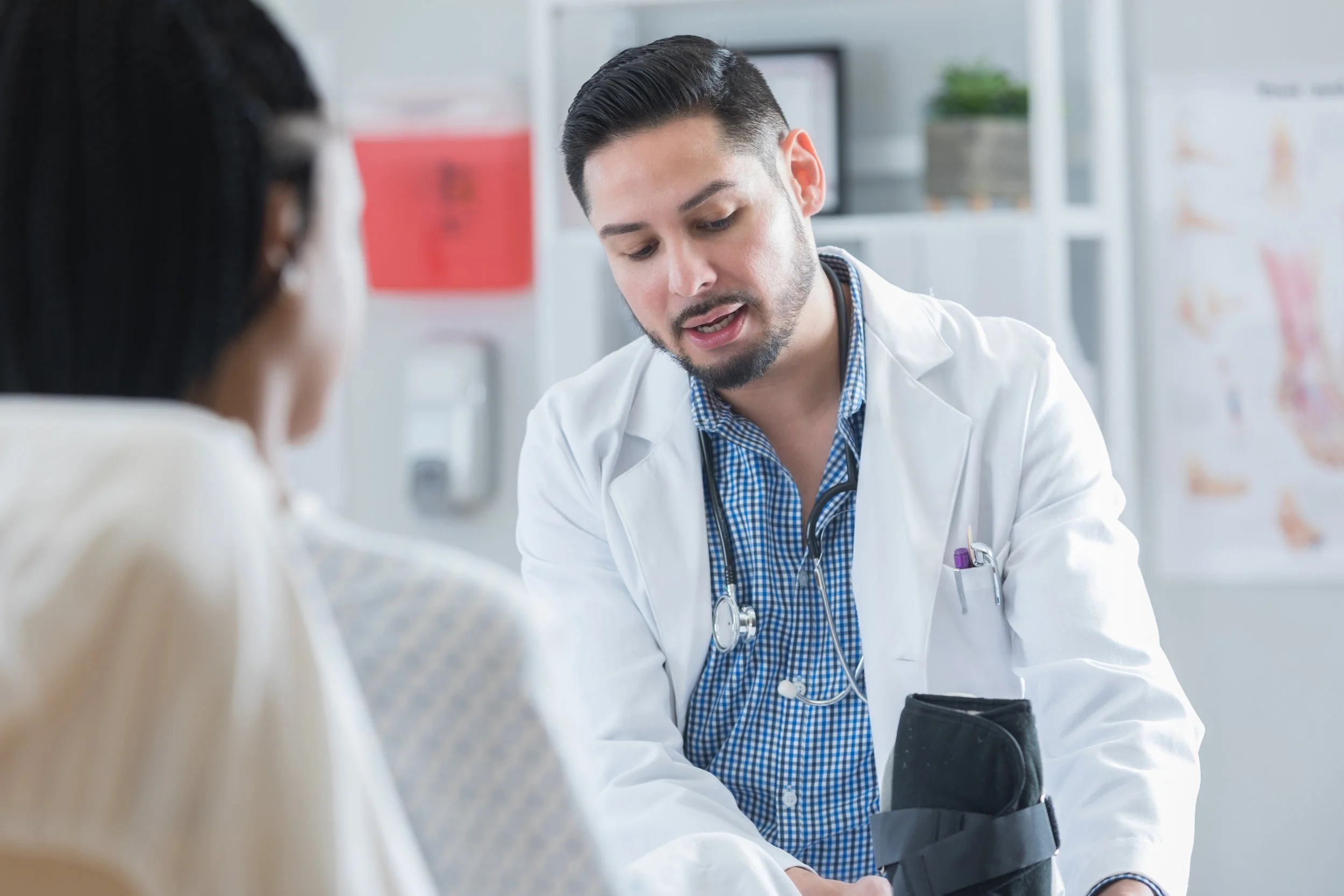 A male doctor in a white coat and blue checkered shirt is checking a patient's blood pressure using a sphygmomanometer in a medical office.