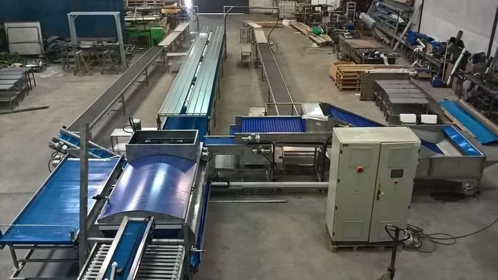 Máquina industrial de processamento, possivelmente de embalagens ou produção, em um ambiente de fábrica com várias áreas de trabalho e equipamentos adicionais ao fundo.