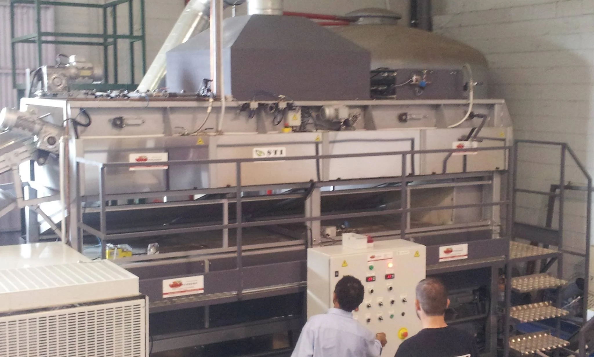 Equipamento industrial de grande porte em um ambiente de fábrica ou laboratório, com pessoas observando e operando a máquina.