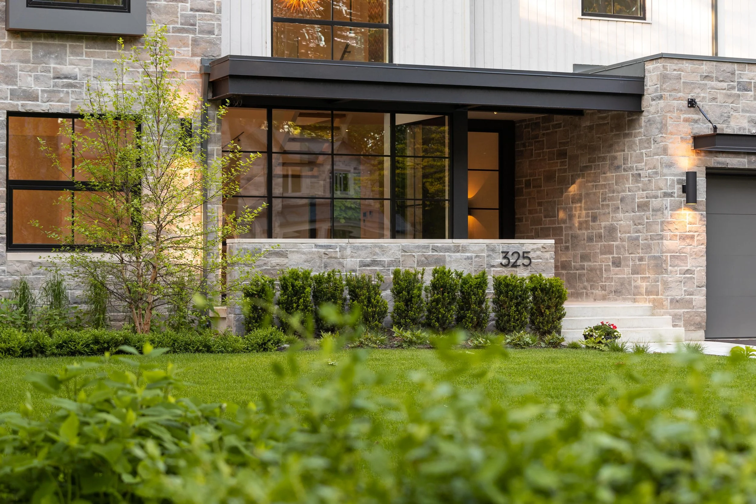 Modern house with brick and stone exterior, large black-framed windows, a small tree and neatly trimmed bushes in front yard, house number 325 on a sign, and a well-maintained lawn.