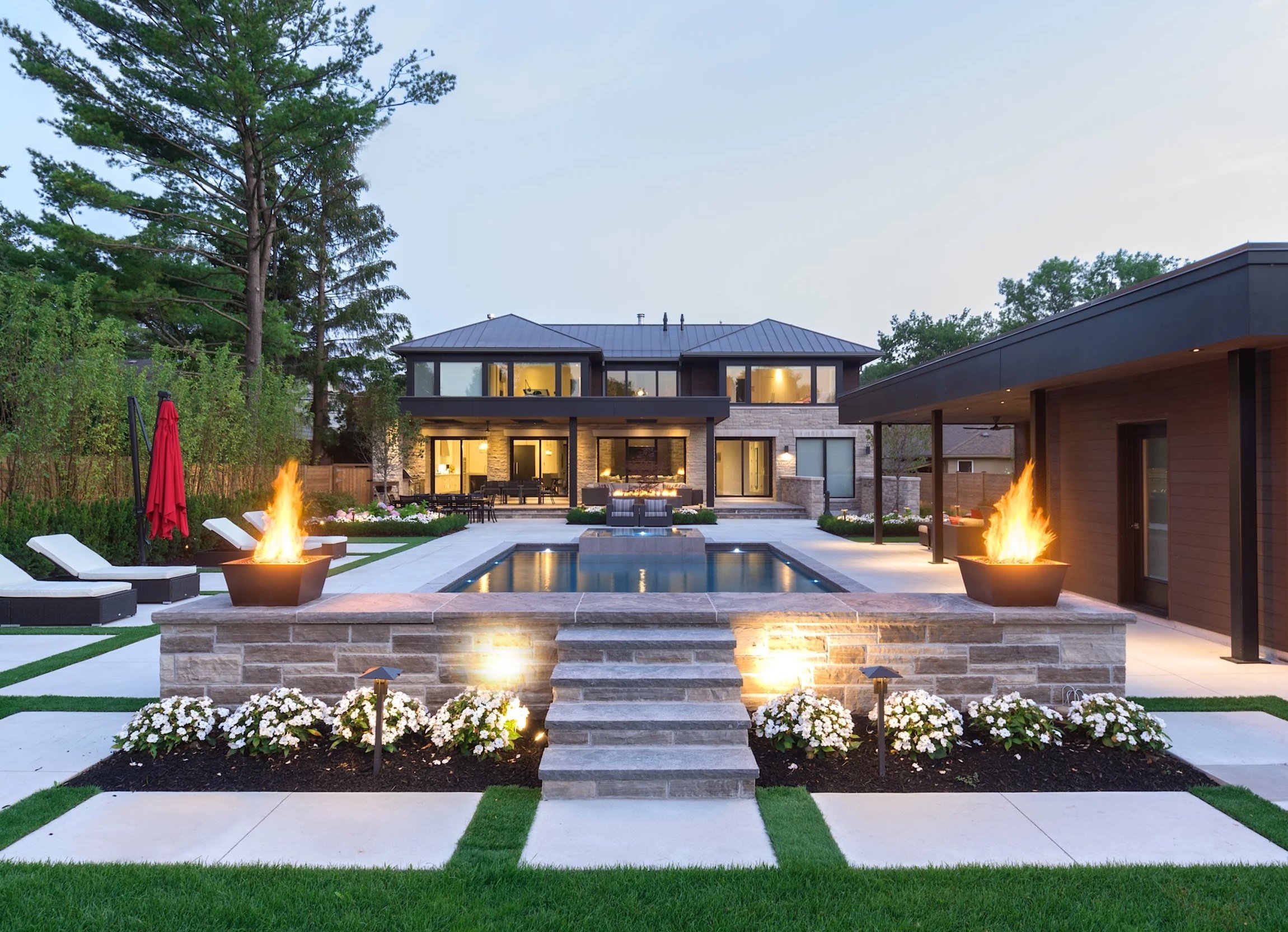 Luxury backyard with in-ground swimming pool, two fire pits, outdoor furniture, flower beds, and a modern two-story house in the background during early evening.