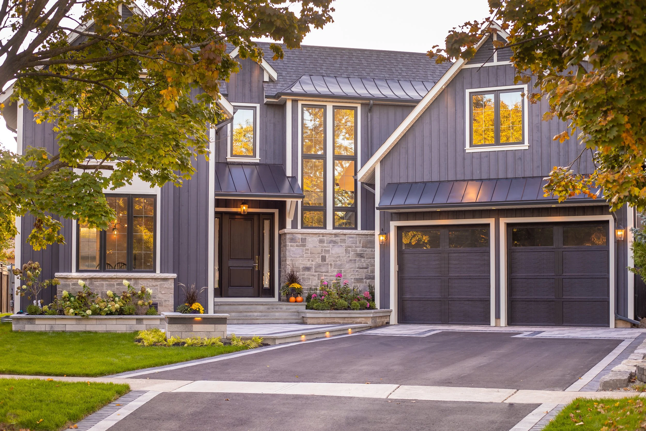 A modern two-story house with dark exterior and large windows, surrounded by a well-manicured lawn, trees, and outdoor lighting in the evening.
