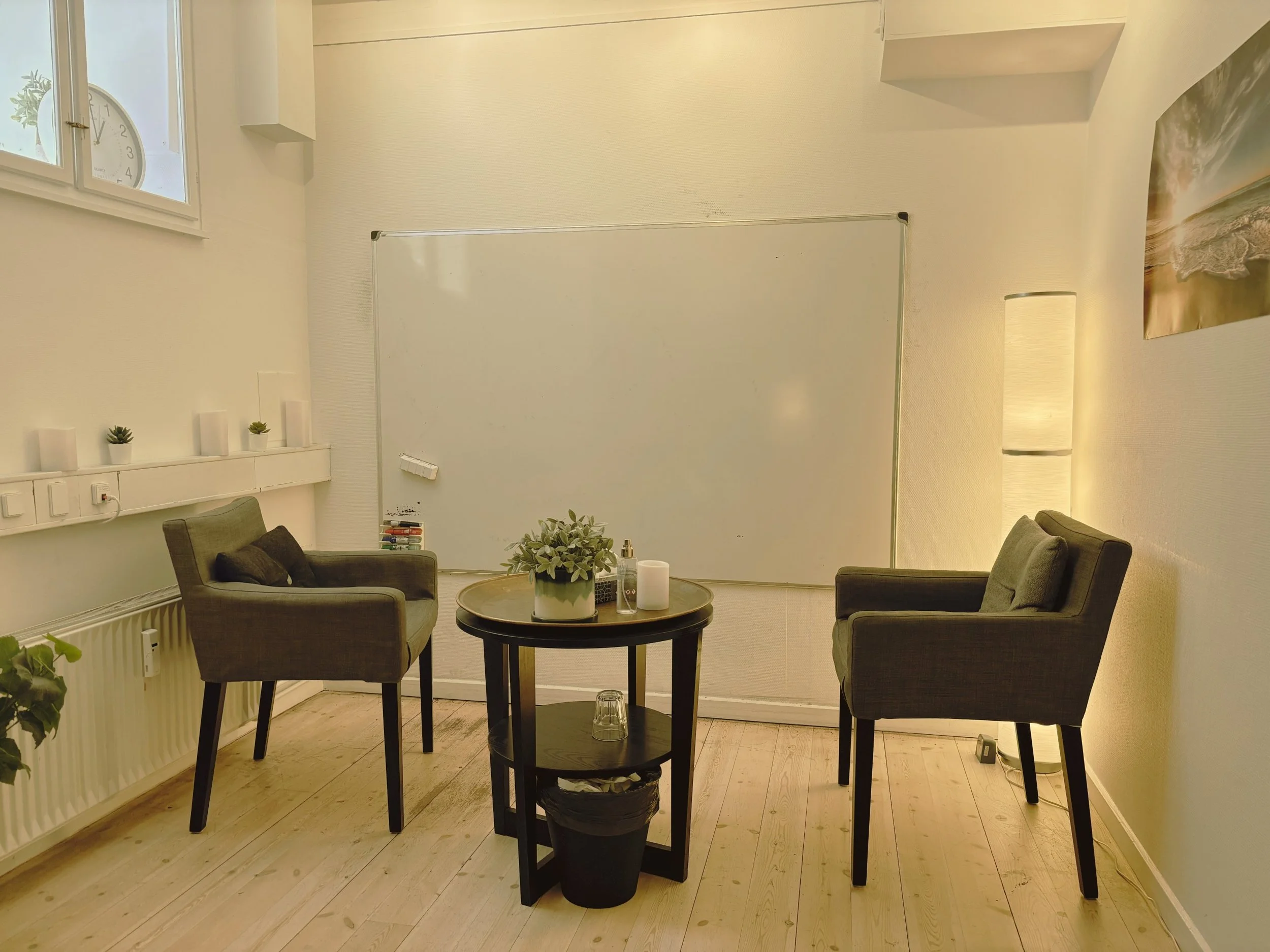 A cozy indoor space with two armchairs, a round table with a plant and candles, a whiteboard on the wall, and a window with a clock showing 1:04. There are small plants and candles on a ledge, a lamp, and a beach poster on the wall.