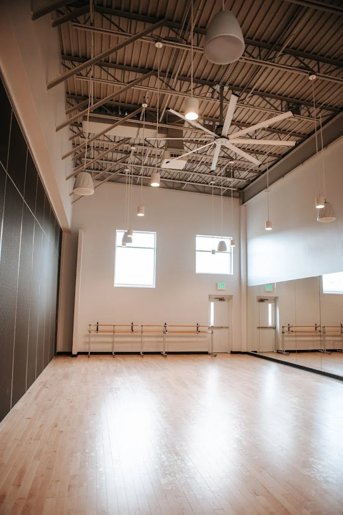 Spacious dance studio with light wood flooring, mirrored walls, ballet barres, and high ceilings with exposed ductwork. Designed for movement, creativity, and performing arts training.
