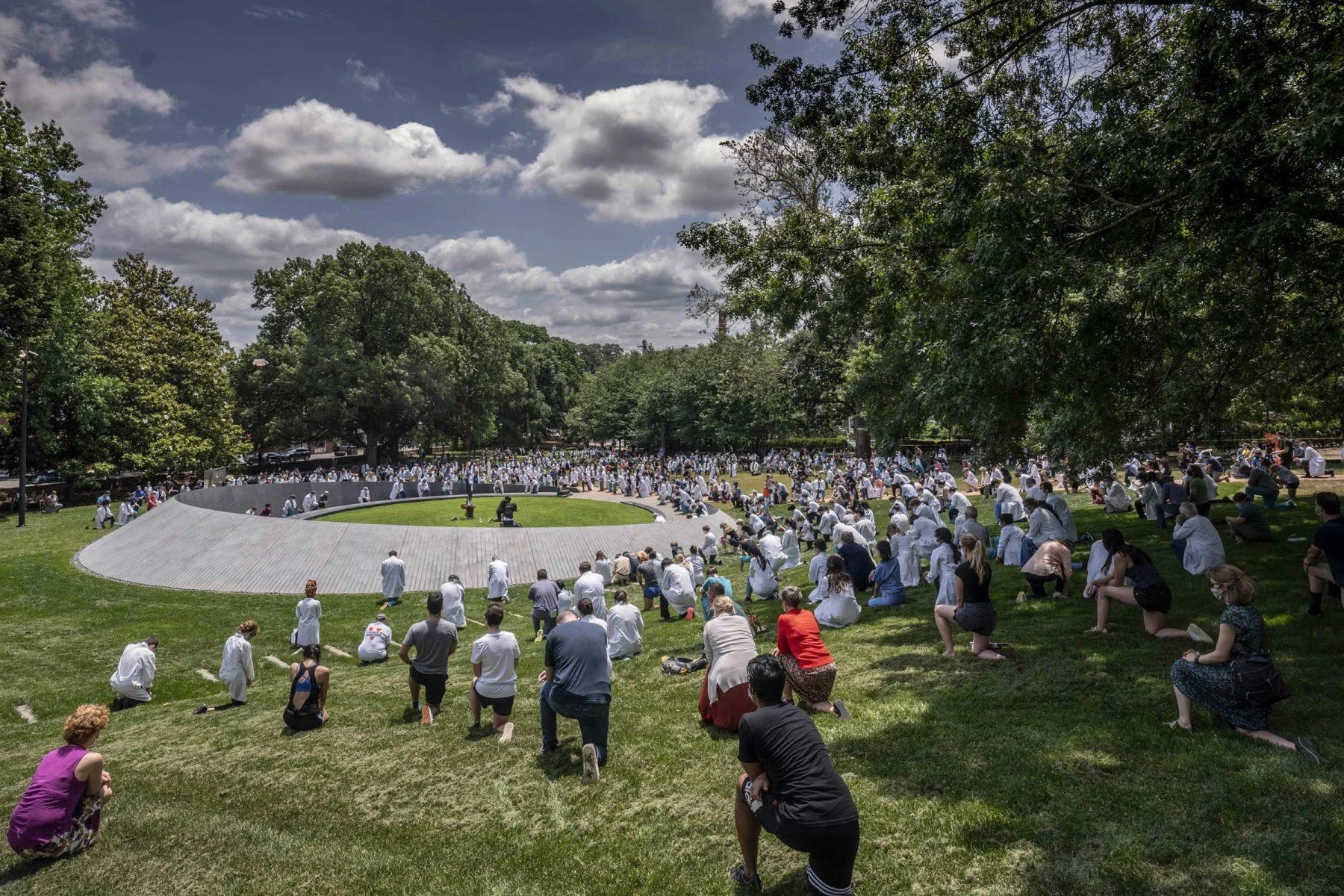Large crowd gathers around a circular monument during a daytime event, many wearing white and kneeling or sitting on the grass. Captures a moment of collective reflection, unity, and public engagement in a space designed for remembrance and community