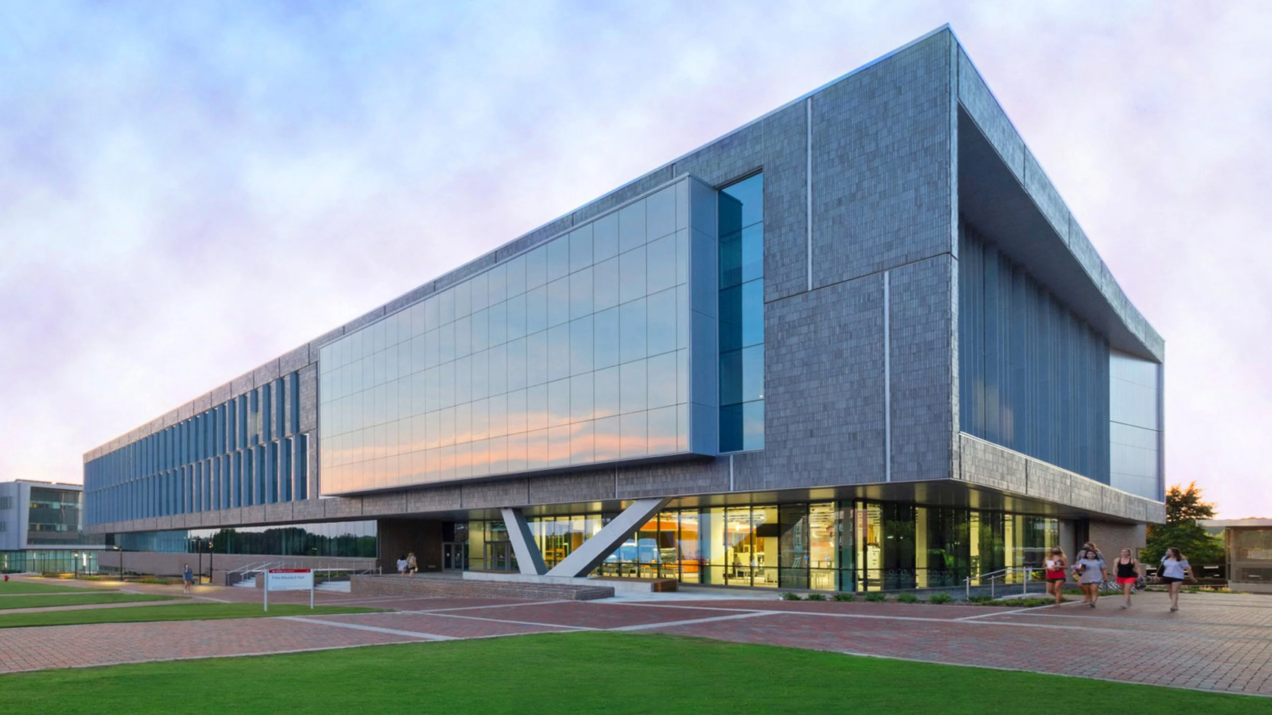 Exterior of a contemporary academic or institutional building with cantilevered glass facade and structural supports. Captures architectural innovation, sunset reflection, and a bold, forward-thinking campus presence.