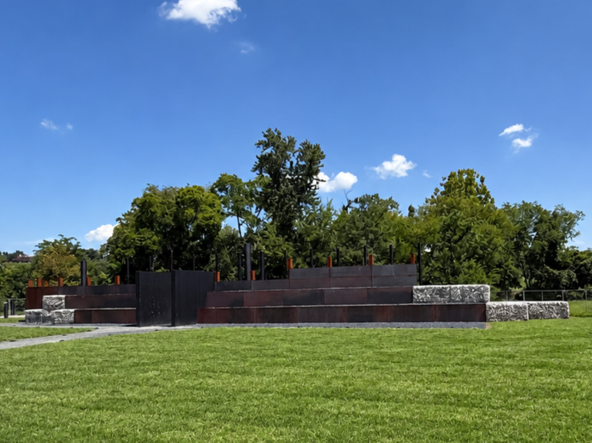 Wide view of a terraced seating structure at a memorial site, with people gathered on rust-colored and stone-faced levels. Emphasizes community engagement, educational gathering, and the blending of natural materials with history.