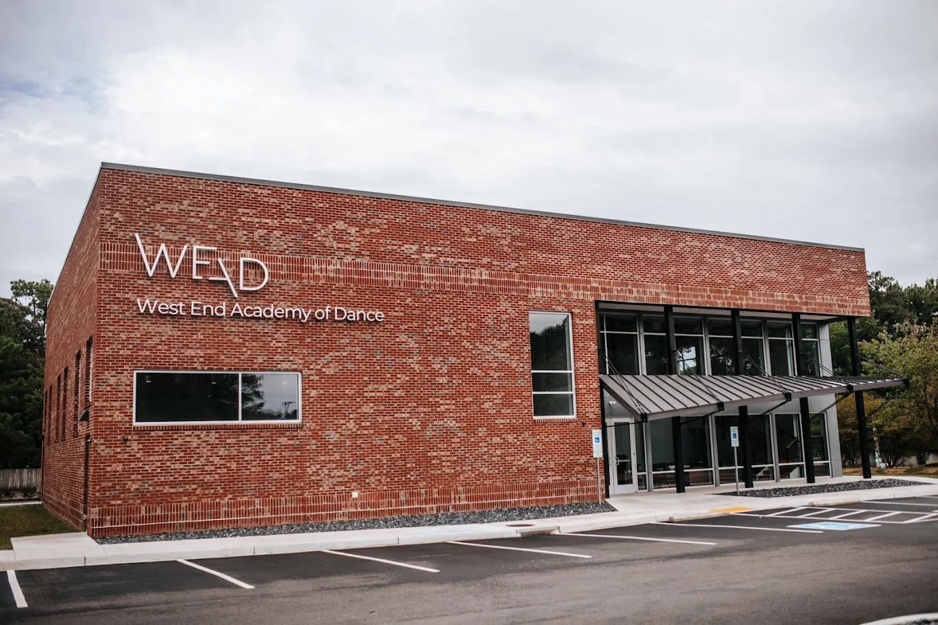 Exterior of the West End Academy of Dance (WEAD), a modern brick building with black-framed windows, a metal awning, and adjacent parking spaces. Highlights clean architectural lines and a welcoming entrance to a performing arts facility.