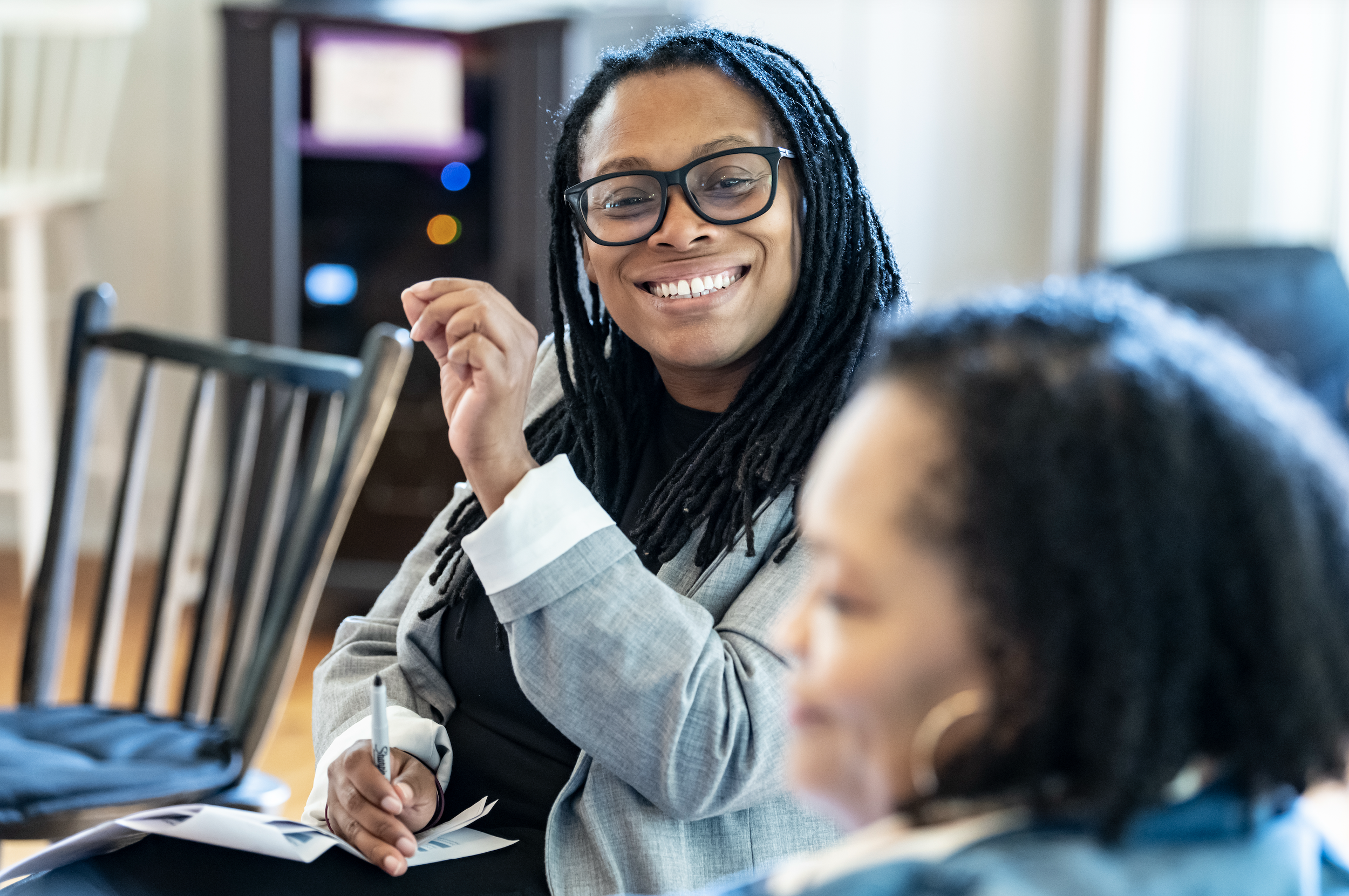 Woman with glasses smiling and taking notes during a meeting or class, with other people present.