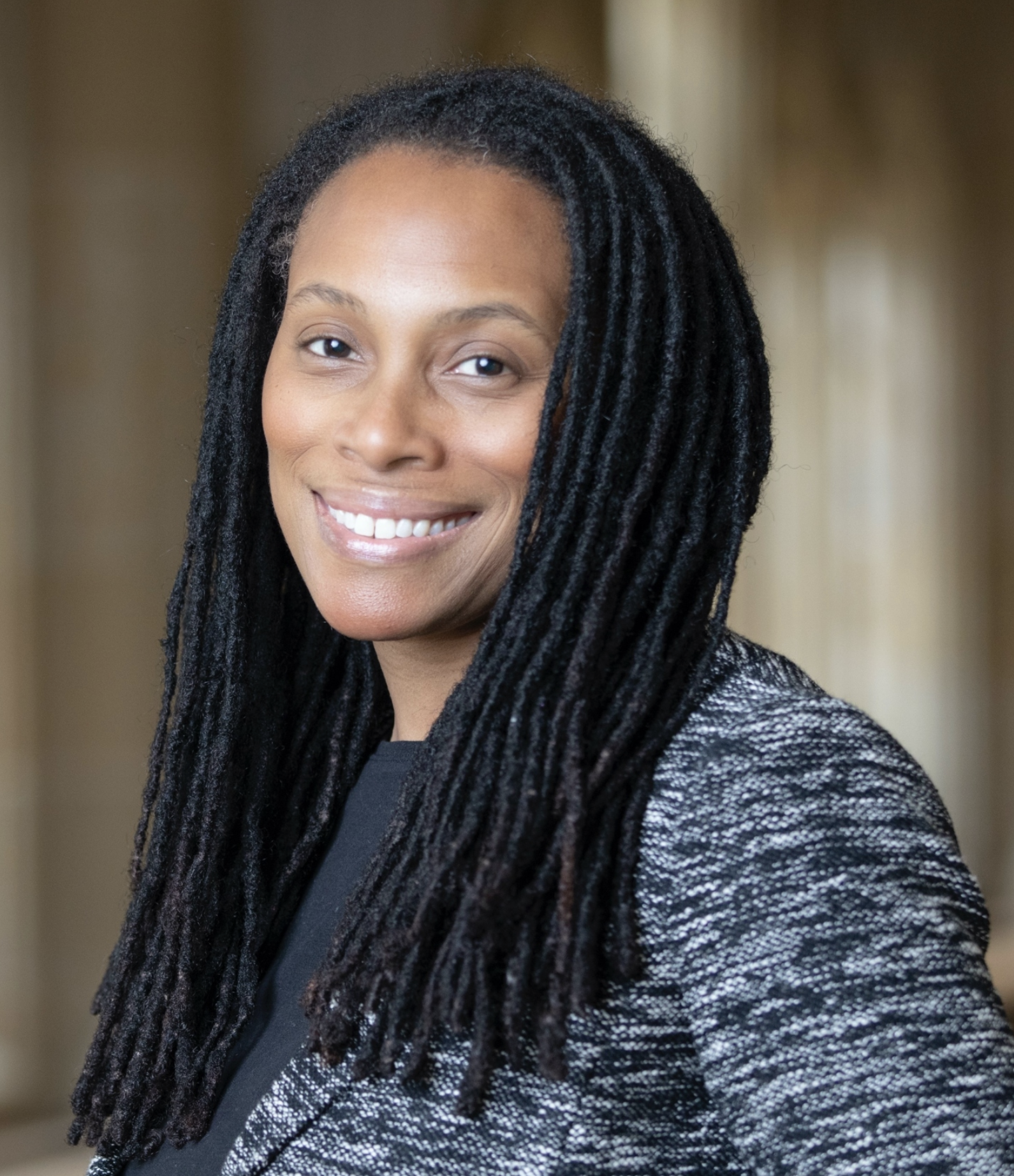 Close-up portrait of a smiling woman with dark skin and long black dreadlocks. She is wearing a dark top and a textured blazer with a blurred indoor background.