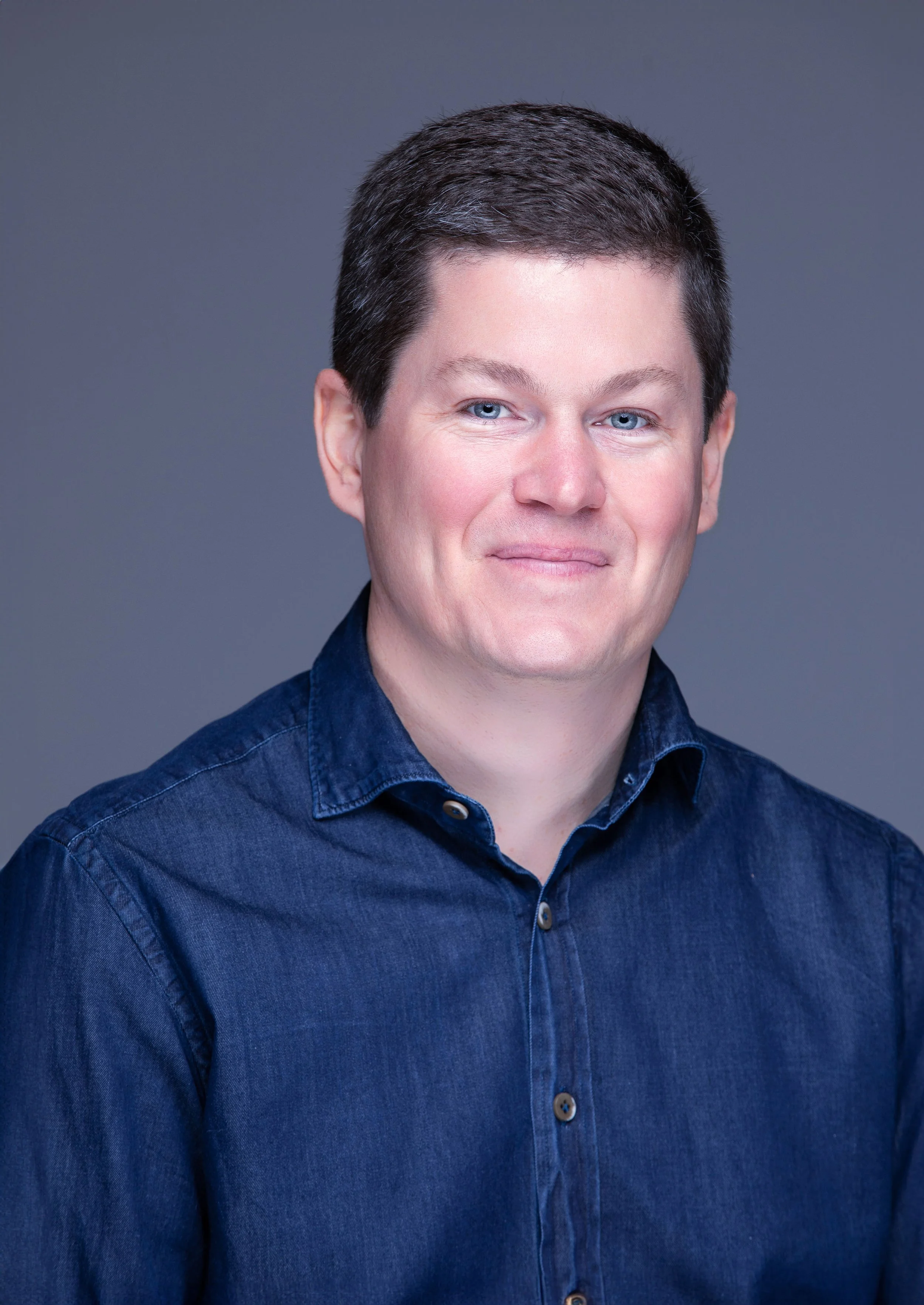 A man with short dark hair and blue eyes wearing a dark blue denim shirt against a neutral gray background.