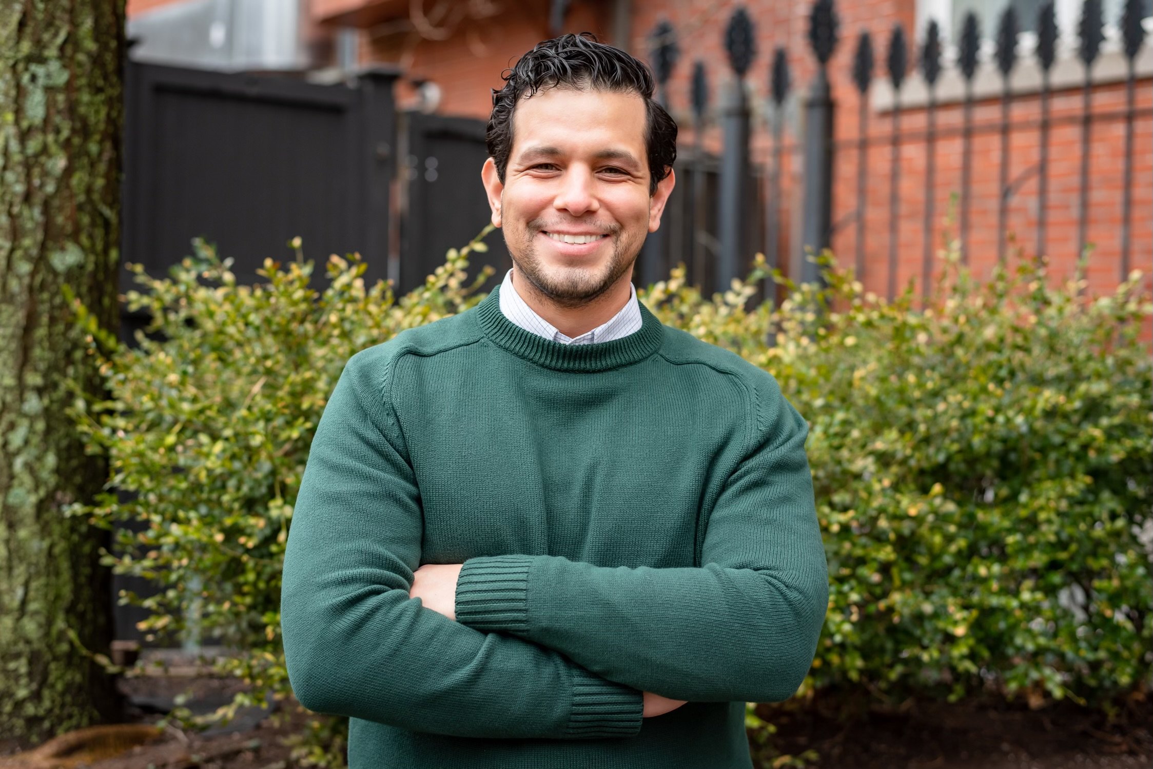 A smiling man with dark curly hair and a beard, wearing a green sweater over a collared shirt, standing outdoors with green bushes and a brick building in the background.