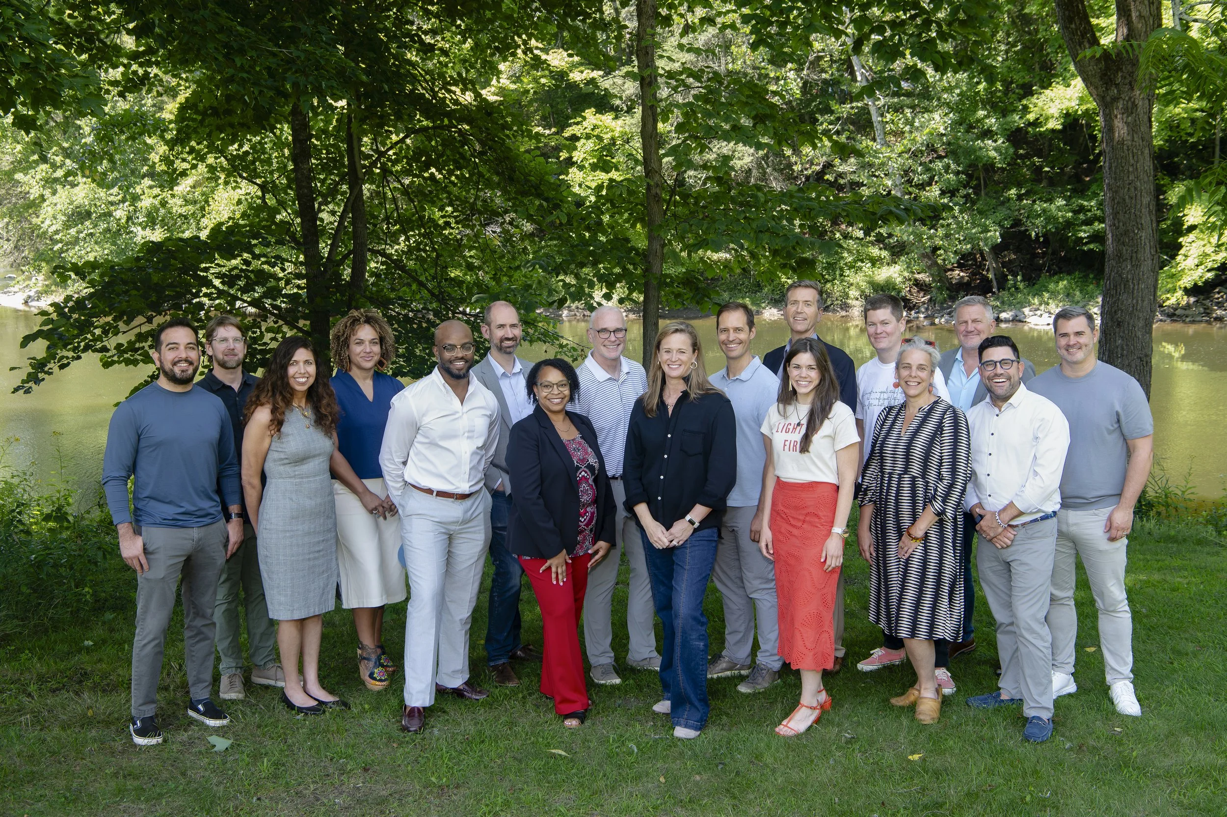 Group of 15 diverse adults posing outdoors near a river, surrounded by trees.
