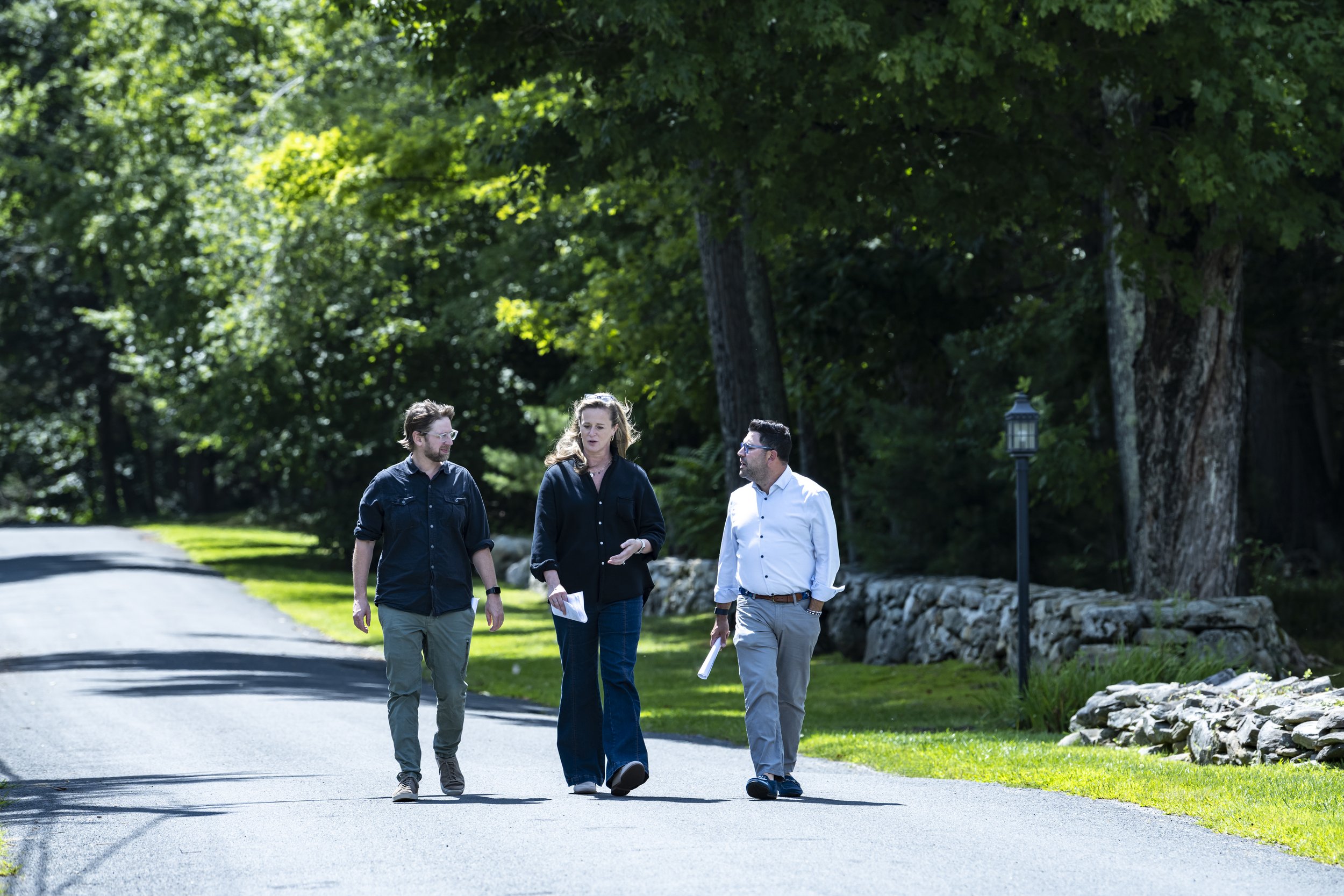 Three people walking outdoors on a pathway through a green park, engaged in conversation. The group includes two men and one woman, dressed in casual business attire, with trees and a lamppost in the background.