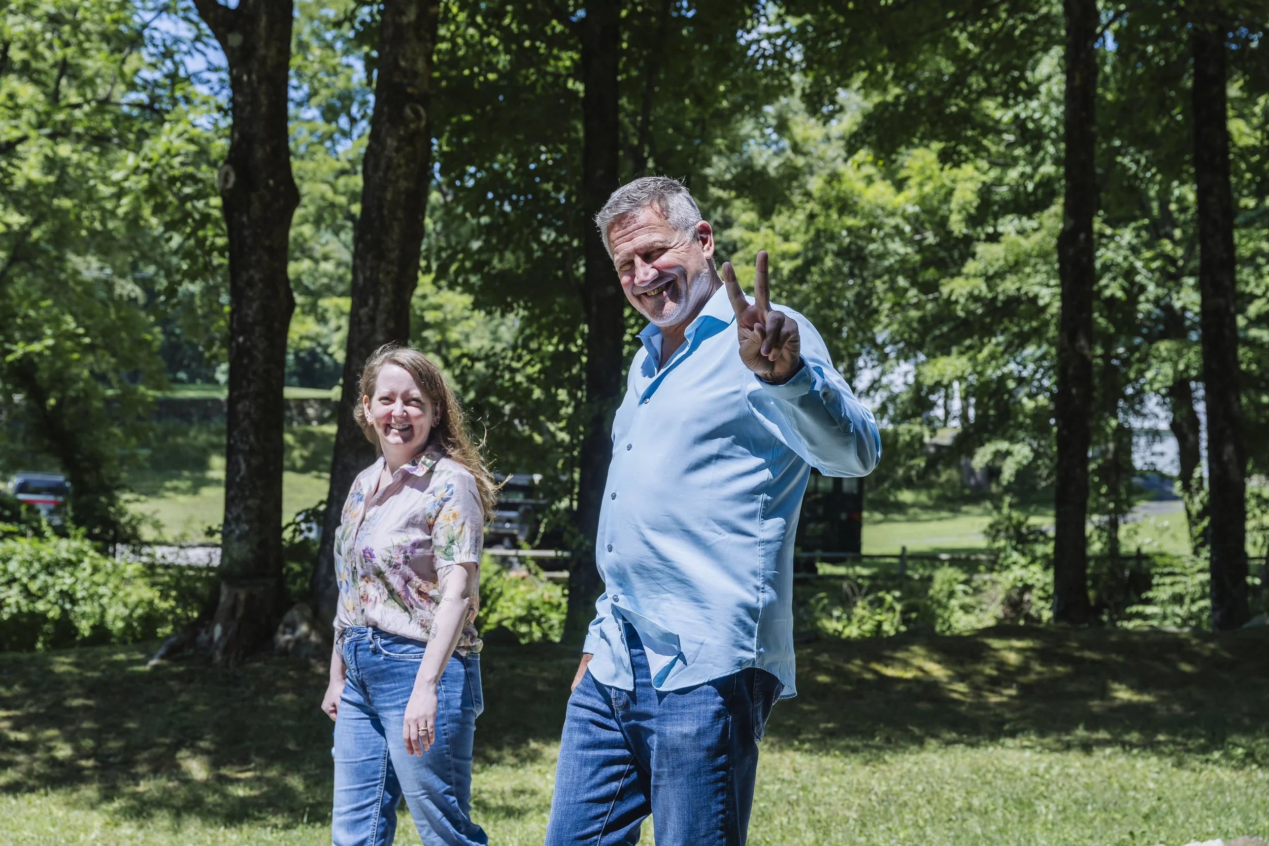 A man and a girl are walking outdoors in a park with tall trees and green grass. The man is smiling and making a peace sign with his fingers, while the girl is smiling.