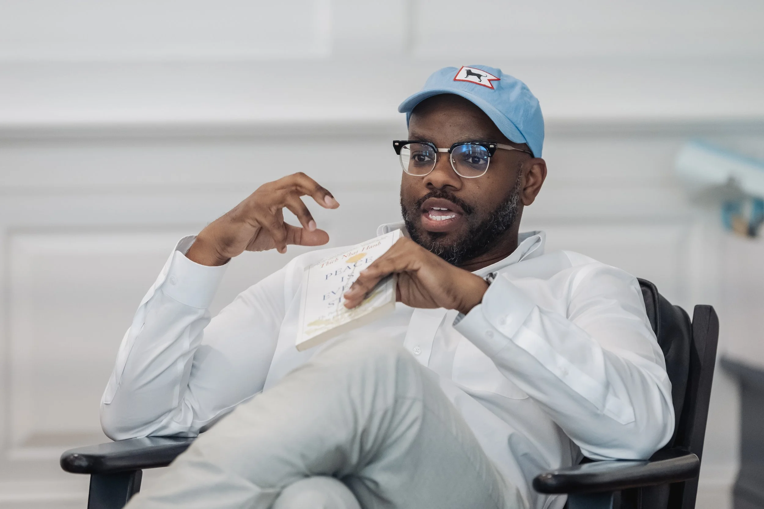 A man wearing a blue cap with a dog patch, glasses, and a white shirt is sitting at a desk in an office, holding a book titled 'Peace' and speaking.