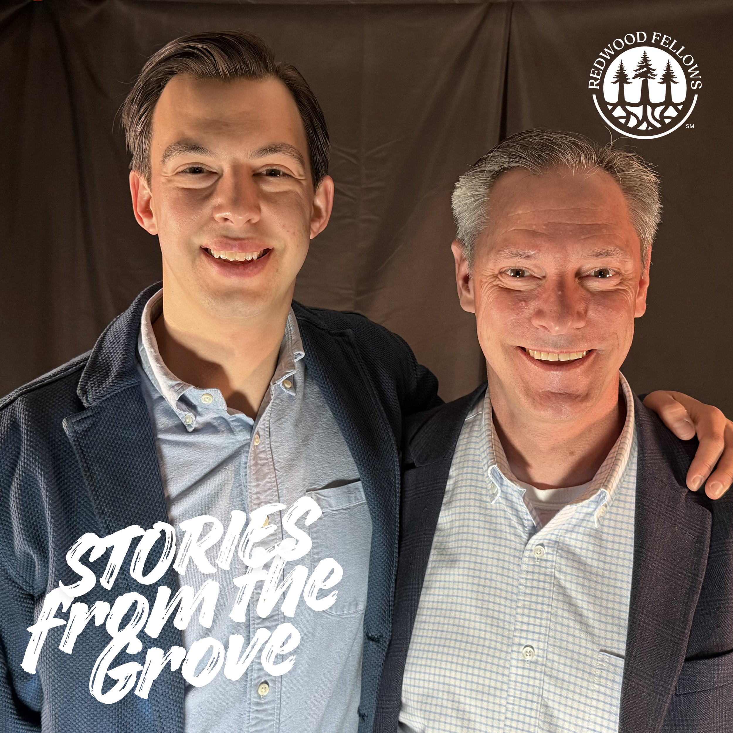 Close-up of two smiling men, one younger with dark hair and the other older with gray hair, standing together against a brown backdrop. The older man is wearing a checkered shirt and the younger man is wearing a dark blazer and a light-colored shirt with a graphic design.